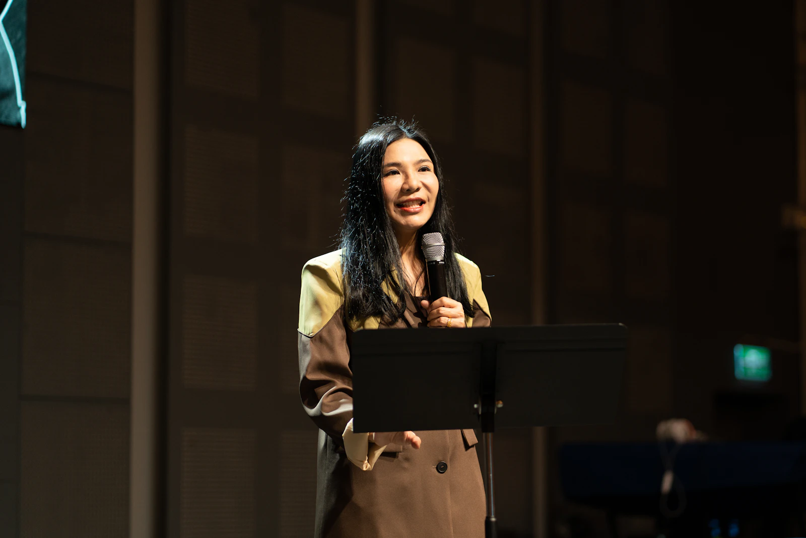 A woman speaking into a microphone on a stage, addressing an audience.
