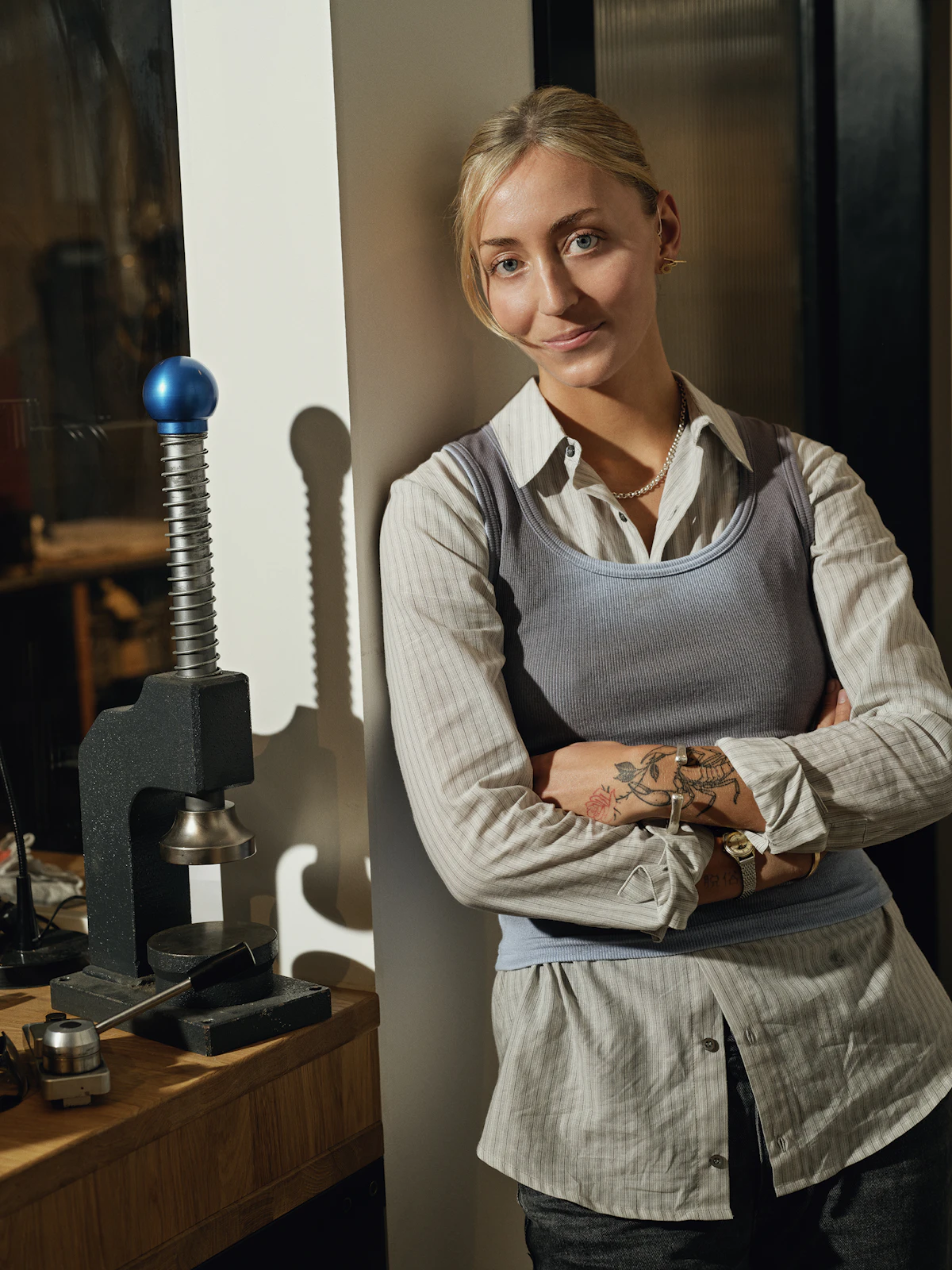 A maker stands at the end of her bench beside a hand press, arms folded, rings on both hands.