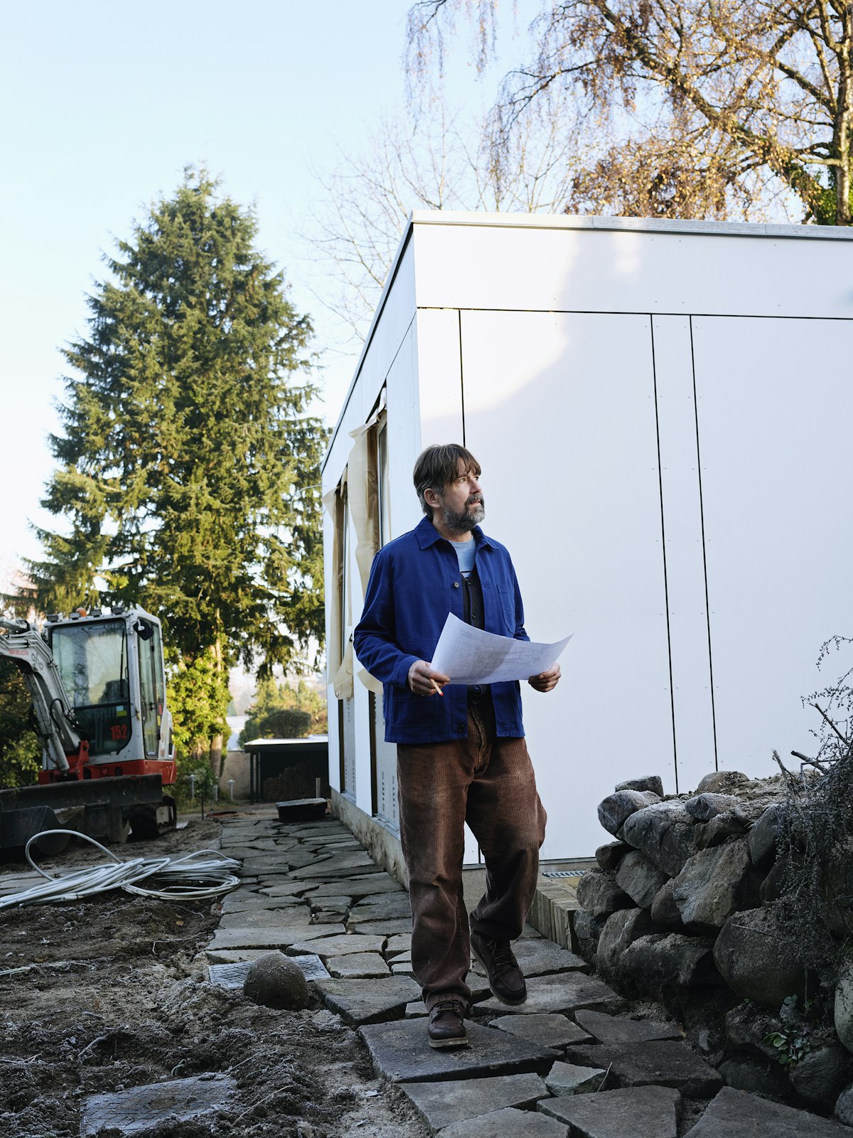 A contractor in a blue jacket holding building plans beside a modern building on a job site
