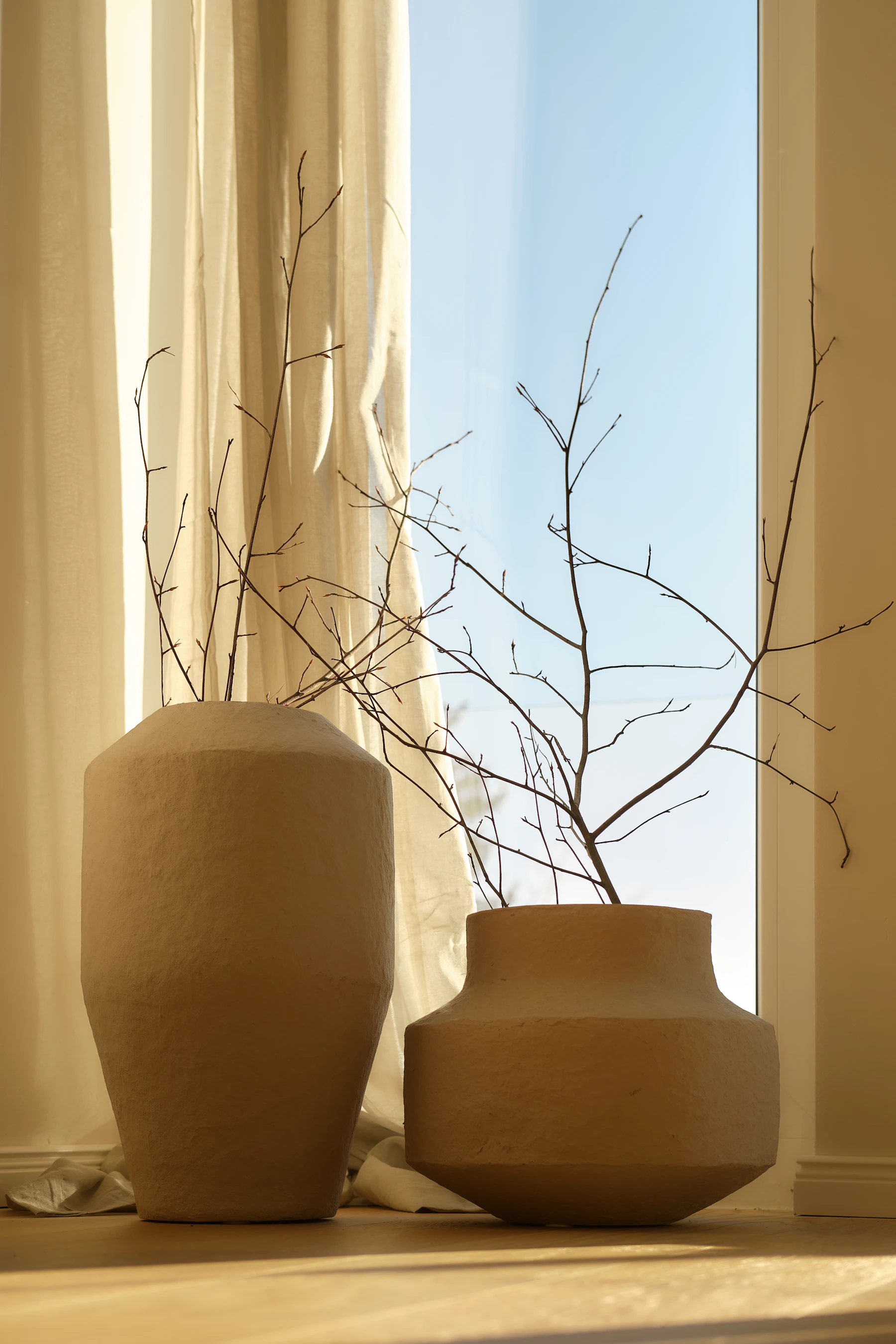 Sculptural neutral ceramic vases with natural branches beside warm linen curtains