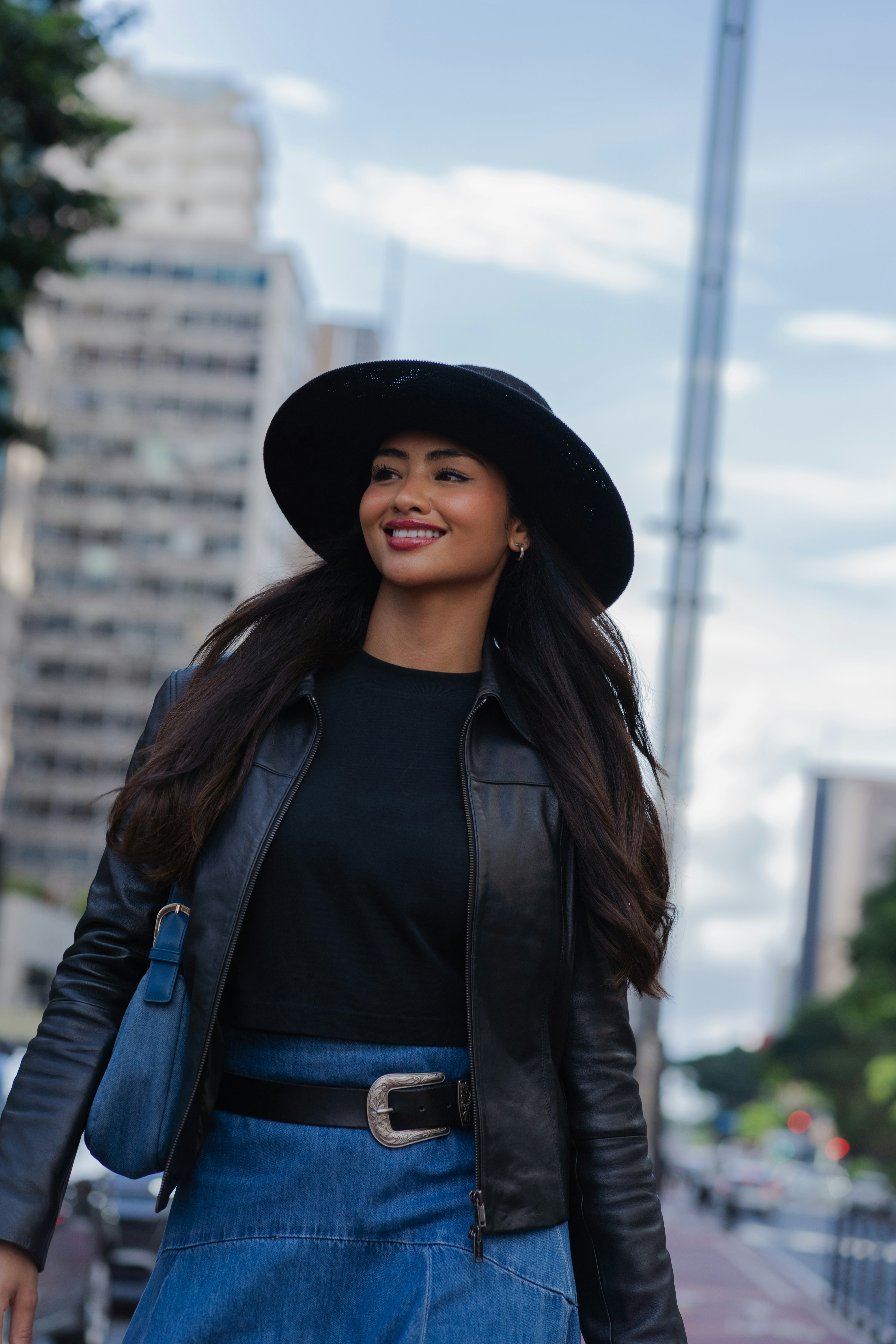 A woman in a hat and leather jacket walks outdoors.