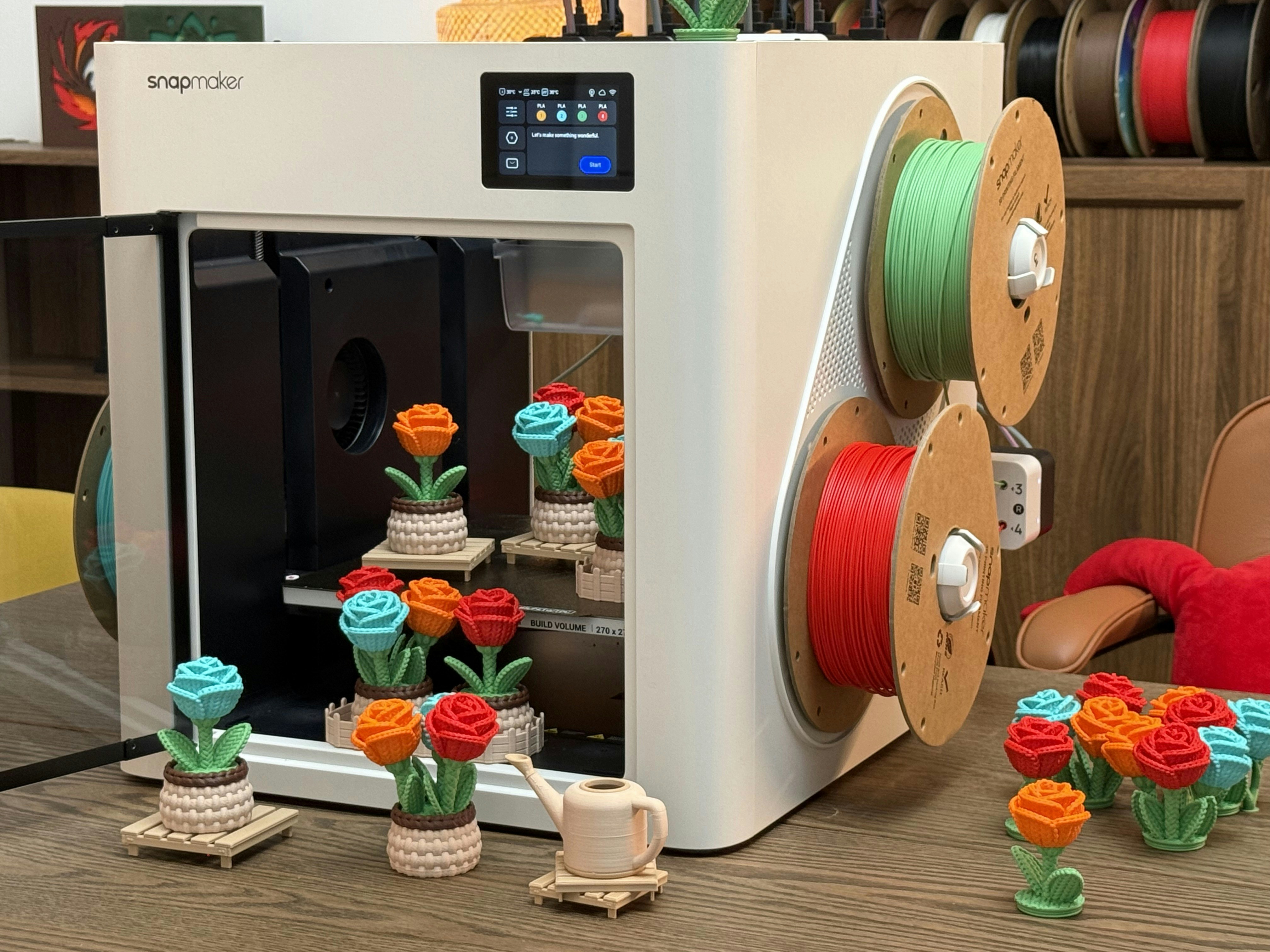 3D model by @flowercat. A Snapmaker 3D printer creating colorful flower models with multiple filament spools in a cozy maker workspace.