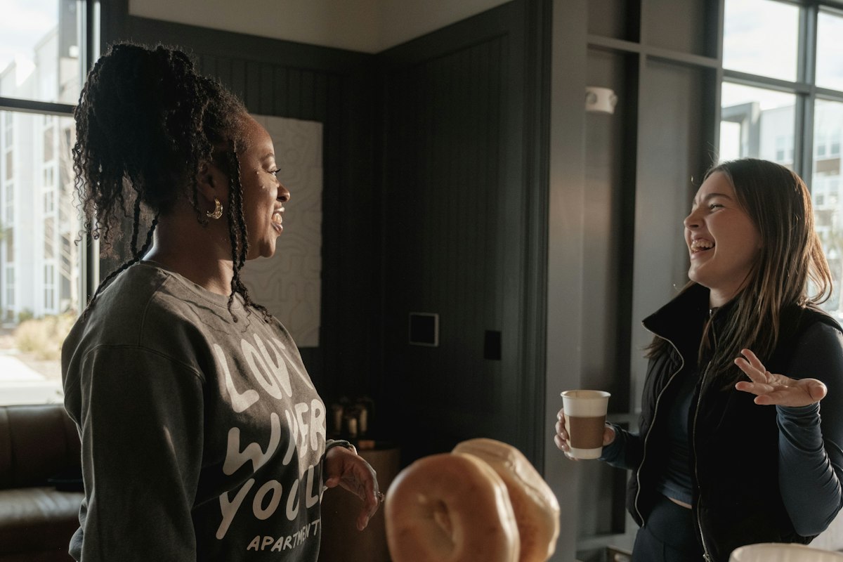 Two women laughing and talking in a sunny room, one holding a coffee cup