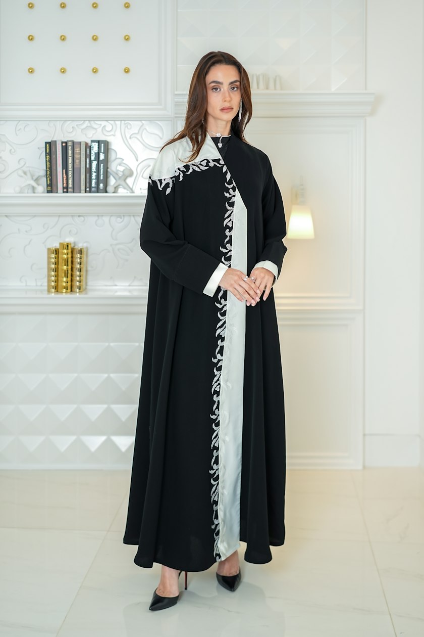 A long, loose black abaya worn open over a white under-dress, full length in frame