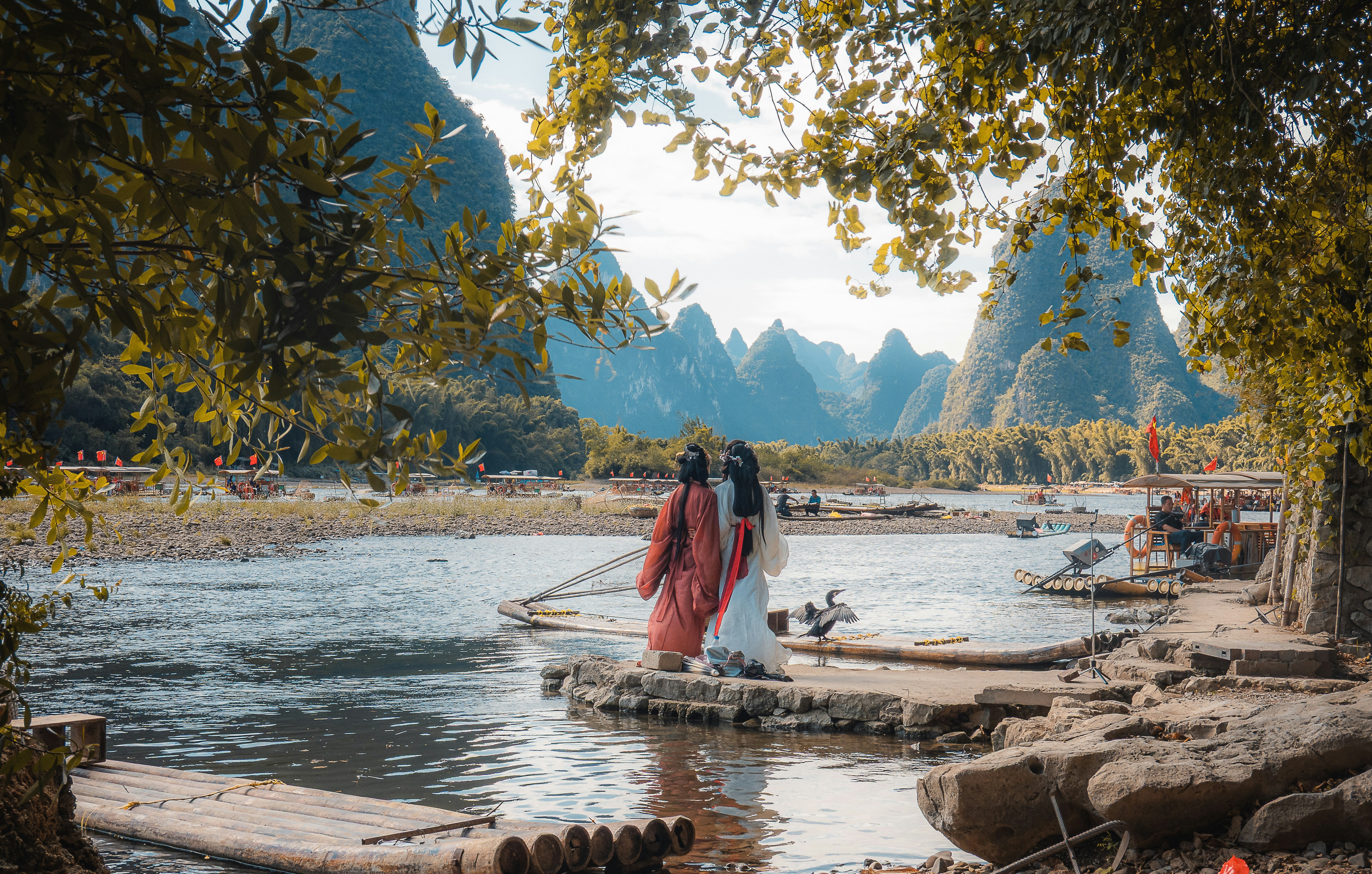Two people in traditional clothing by a river with mountains.