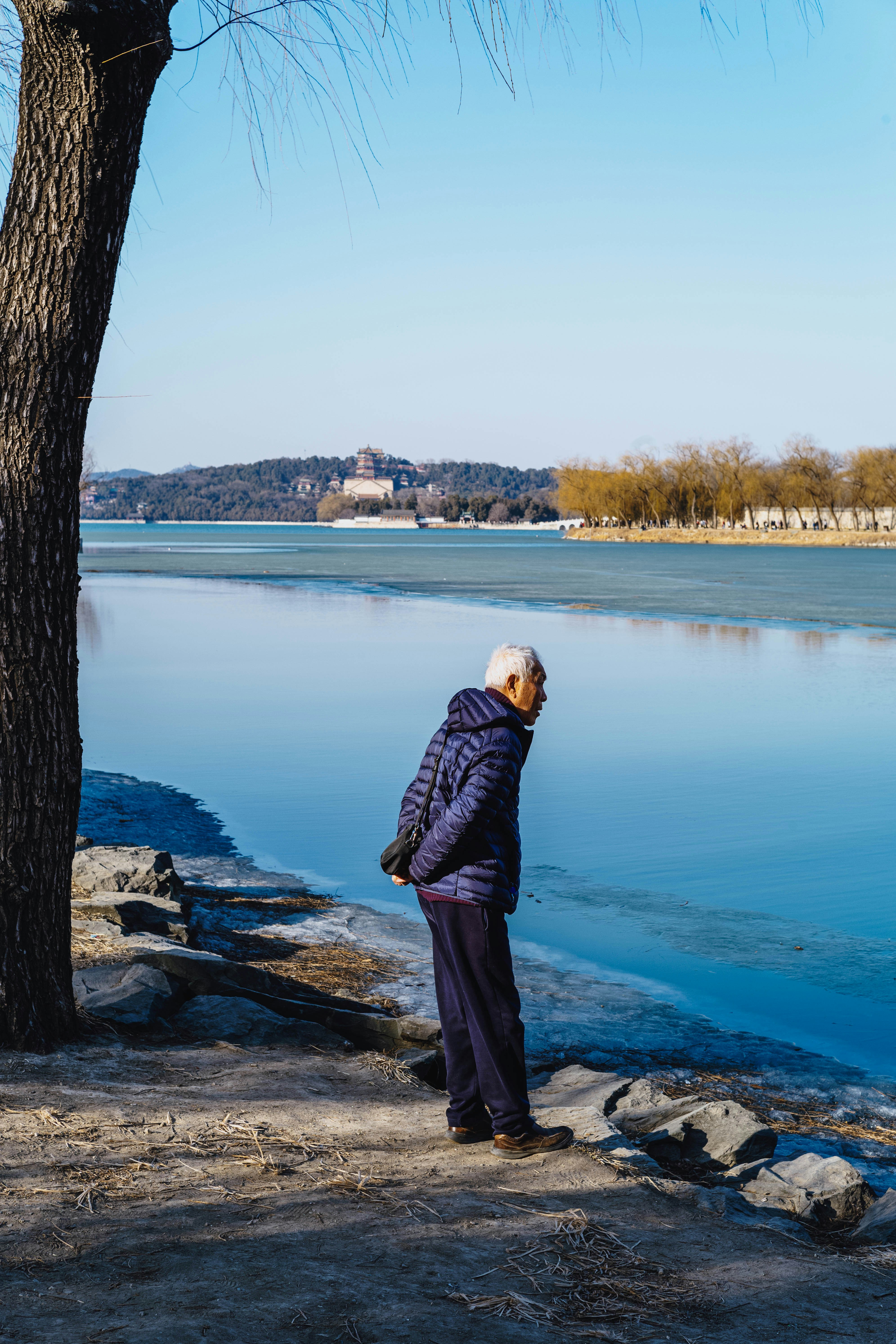 Un vieil homme se tient près d’un lac gelé avec des bâtiments éloignés.