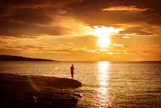 A lone figure stands on a rocky shore at sunset.