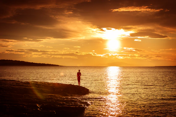 A lone figure stands on a rocky shore at sunset.