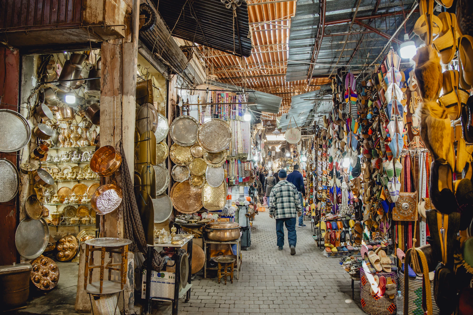 Travelers walk through a vibrant Marrakech souk lined with metalwork, textiles, and slippers