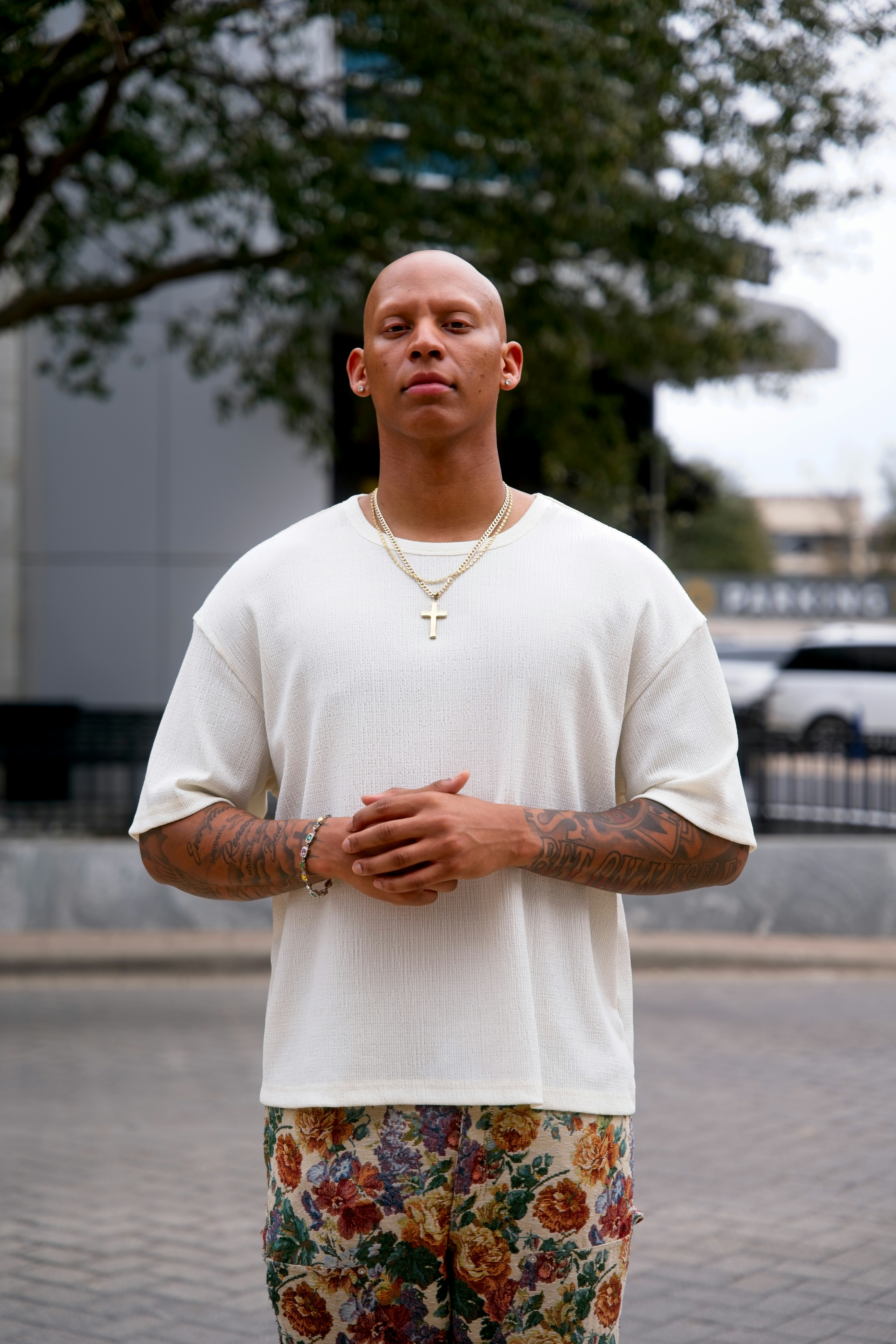 Homem careca com tatuagens, usando calças florais e camisa branca.