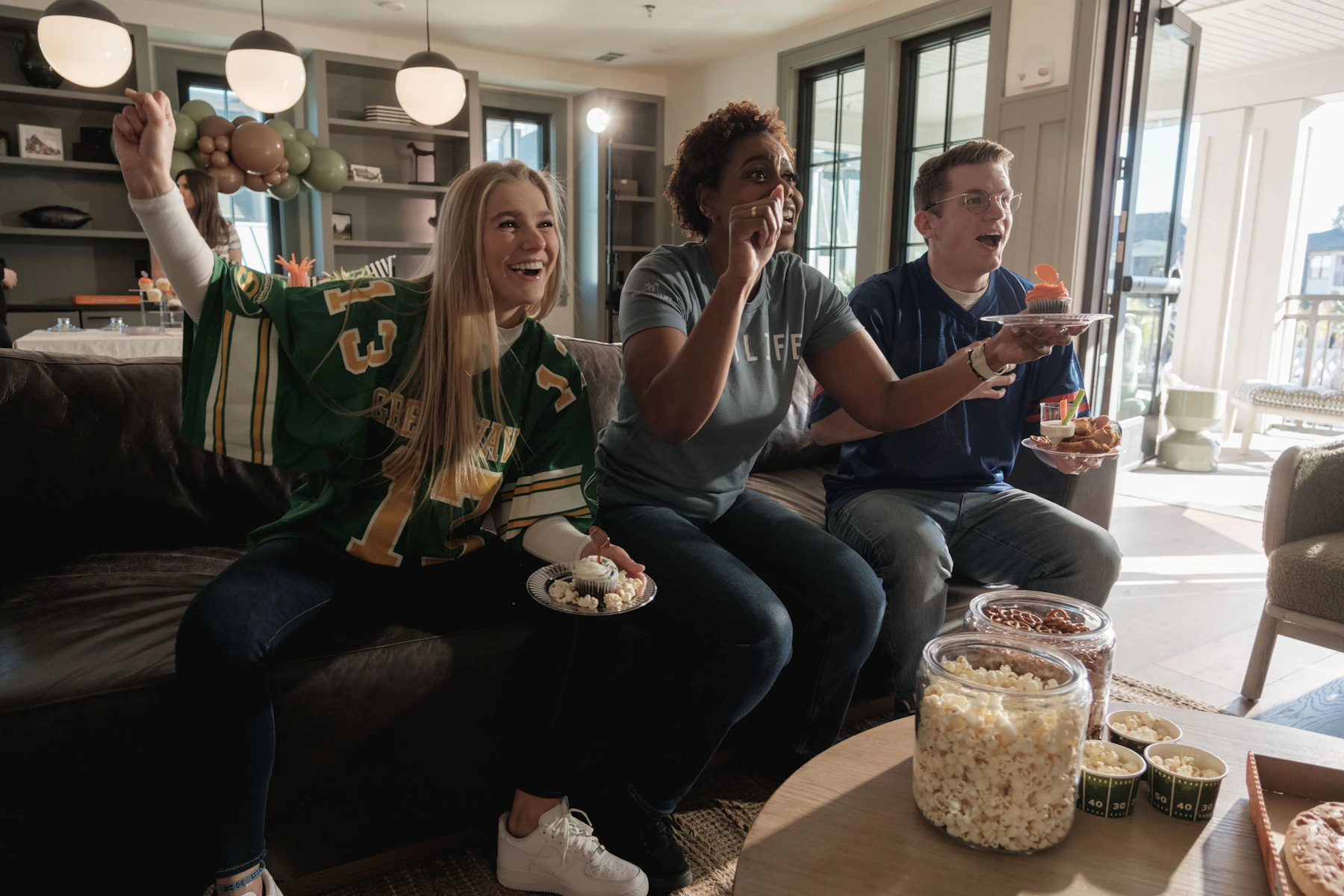 Friends in football jerseys cheering on a sofa with popcorn