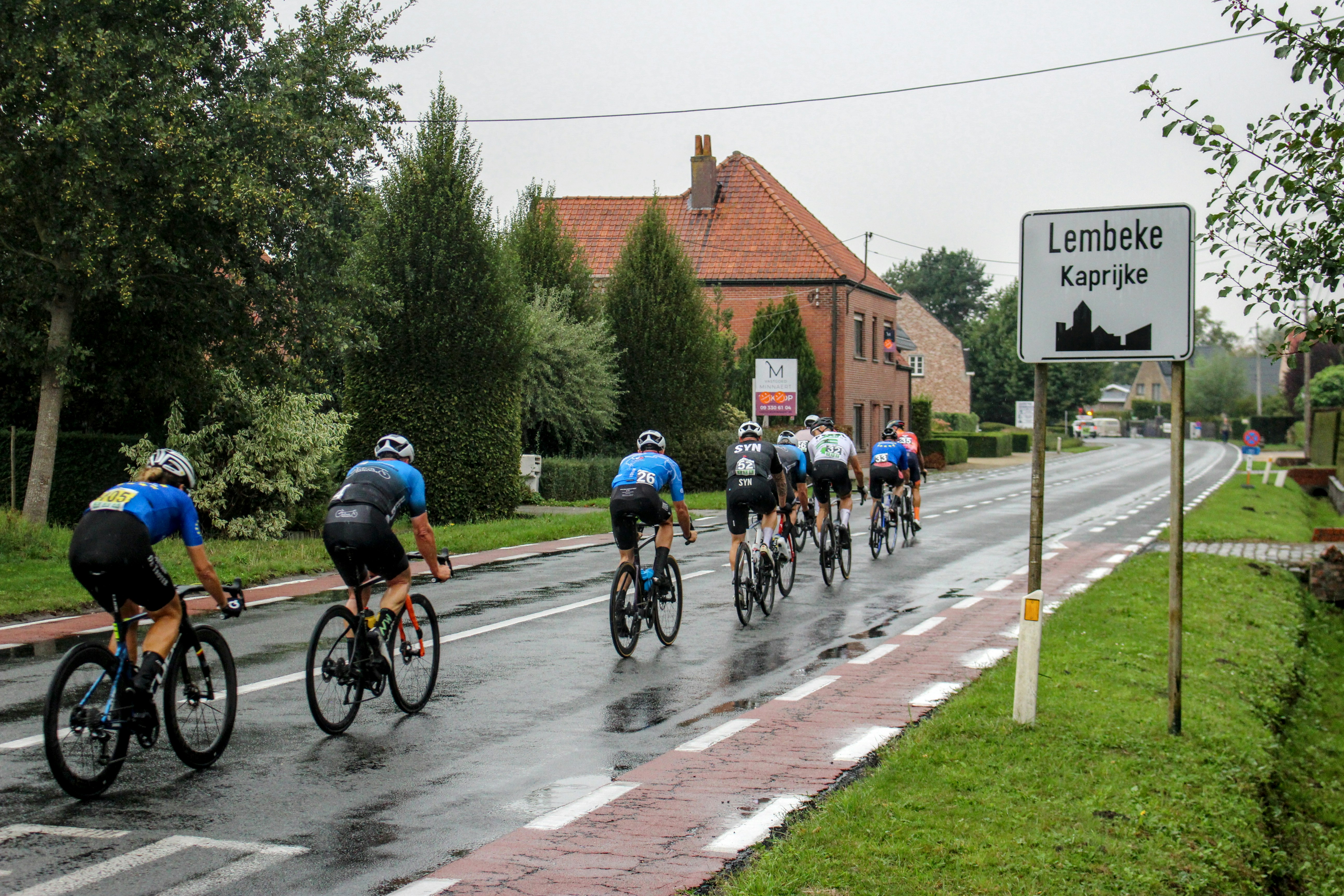 Cyclists ride on a wet road past a village sign.