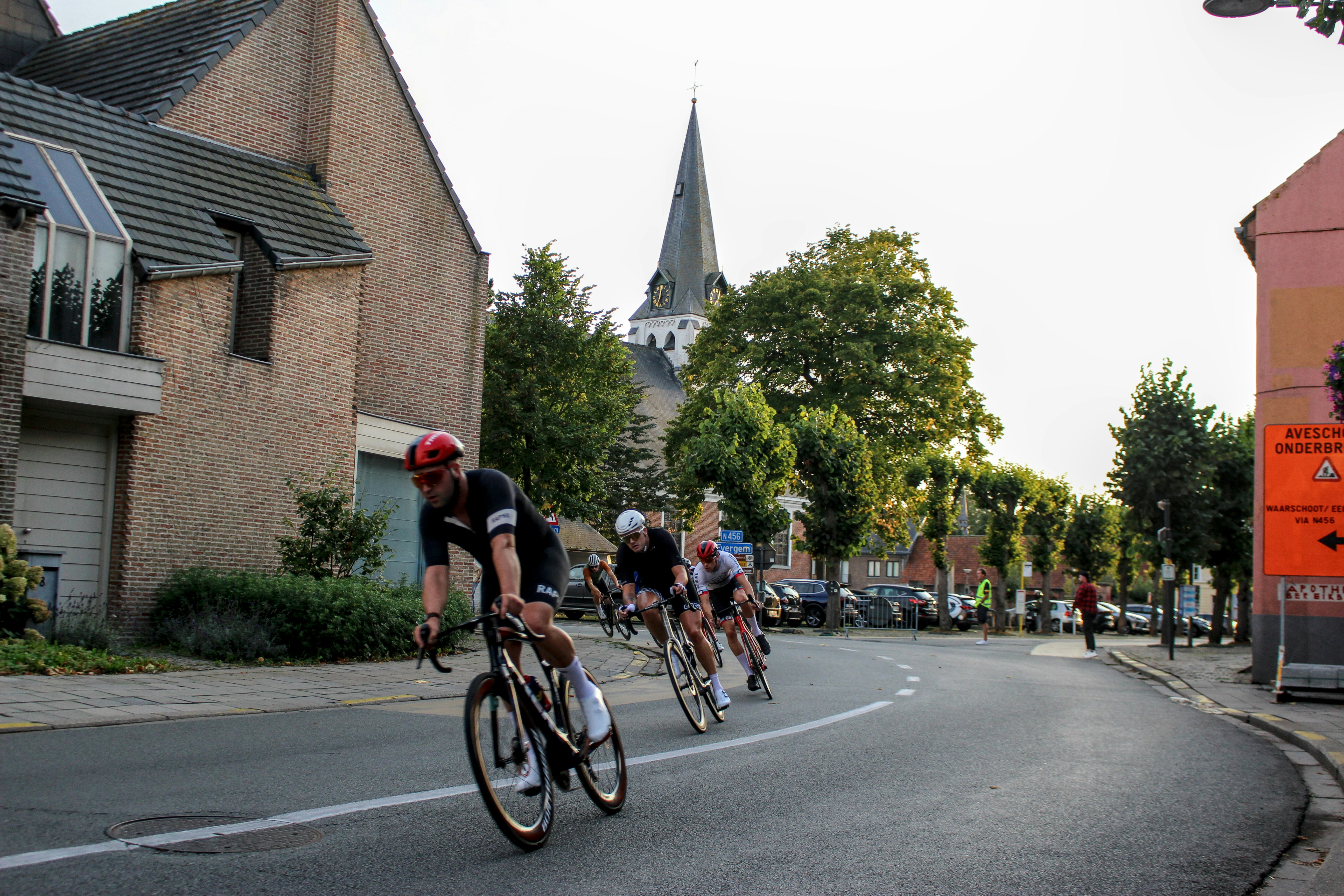 Cyclists race down a street with buildings and trees.