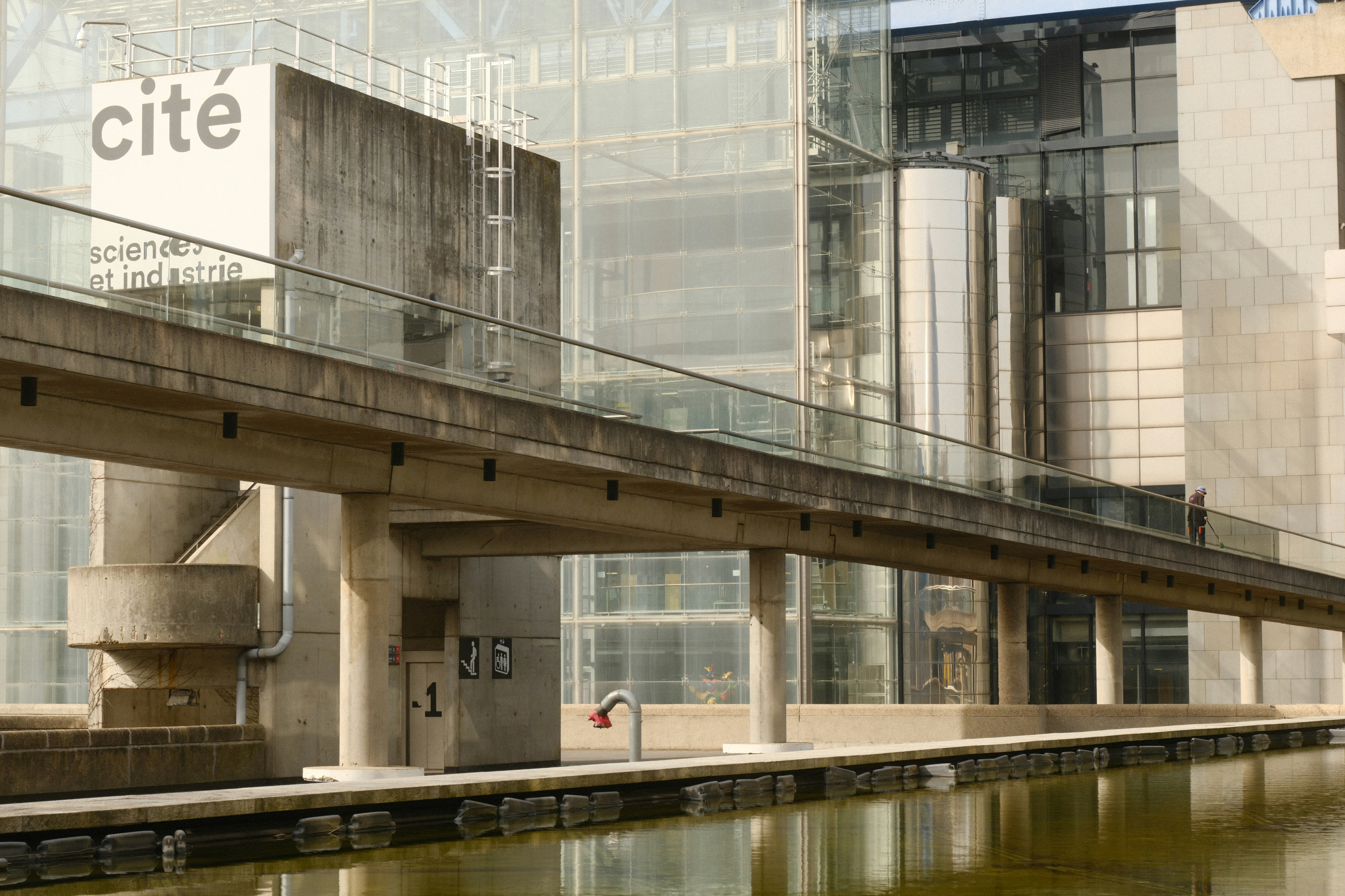 Modern concrete walkway over water with glass buildings.