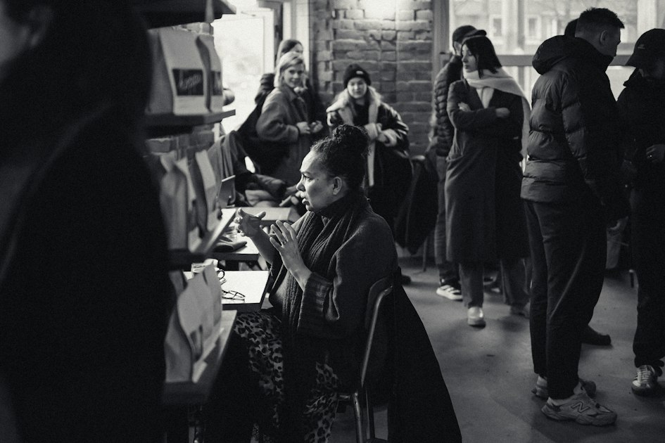 Black and white photograph of a woman mid-gesture explaining something to a group in a crowded room, with other people listening around her