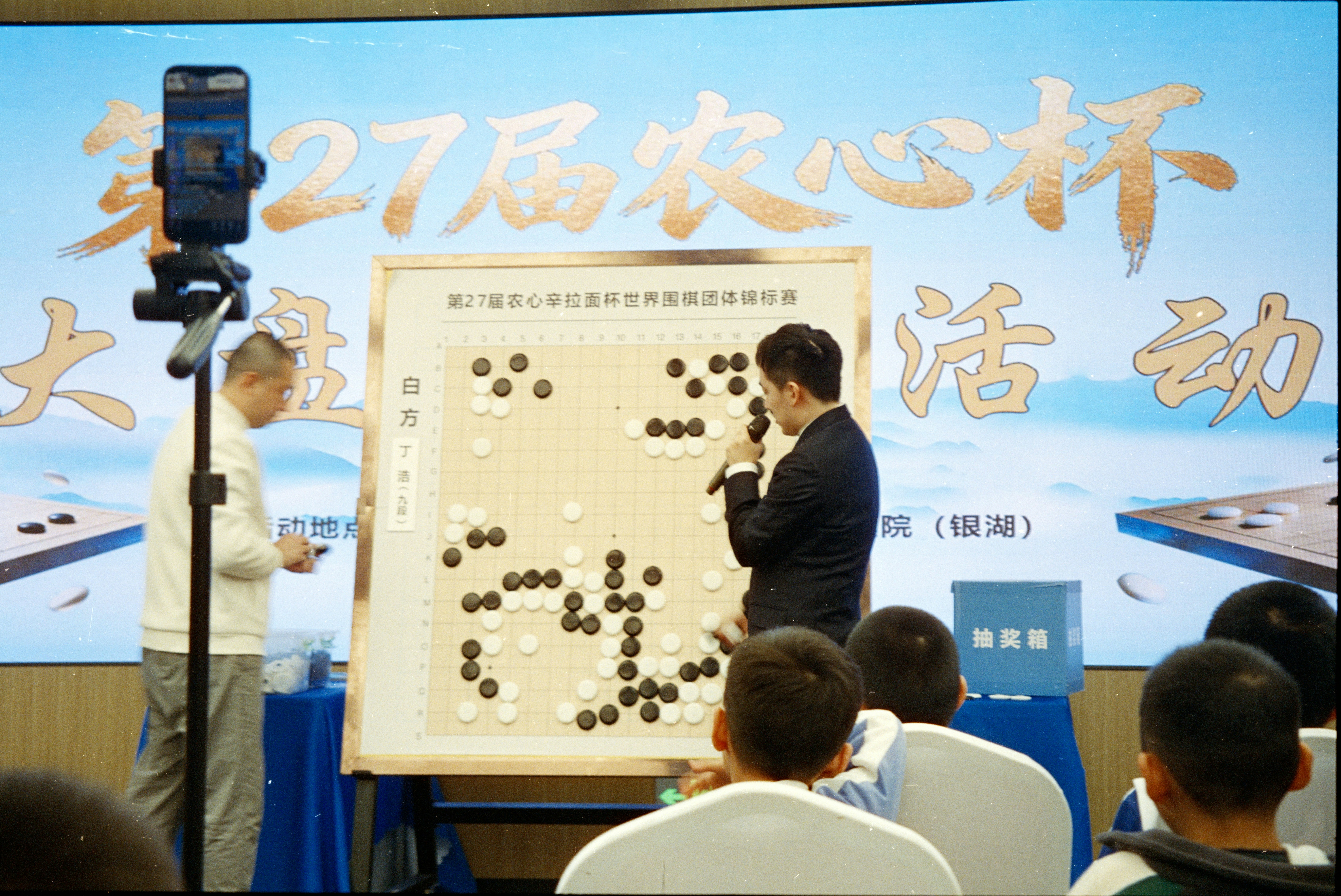 大型白色棋盤前，一名男性講解員正在分析棋局，台下有觀眾專注聆聽，背景有第27屆農心杯比賽字樣