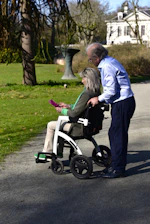 Elderly couple walking in a park with a wheelchair.