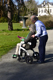 Elderly couple walking in a park with a wheelchair.