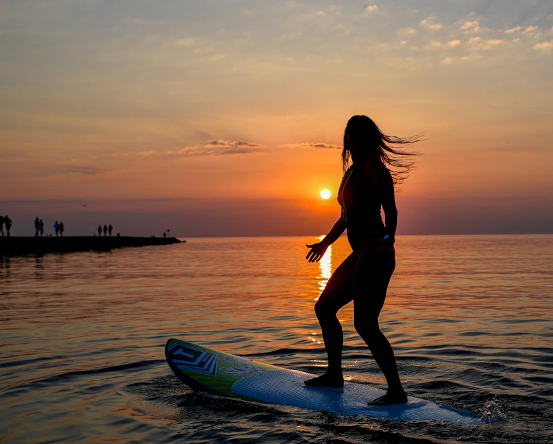 A woman standing on her surfboard in the ocean as the sun sets on the horizon