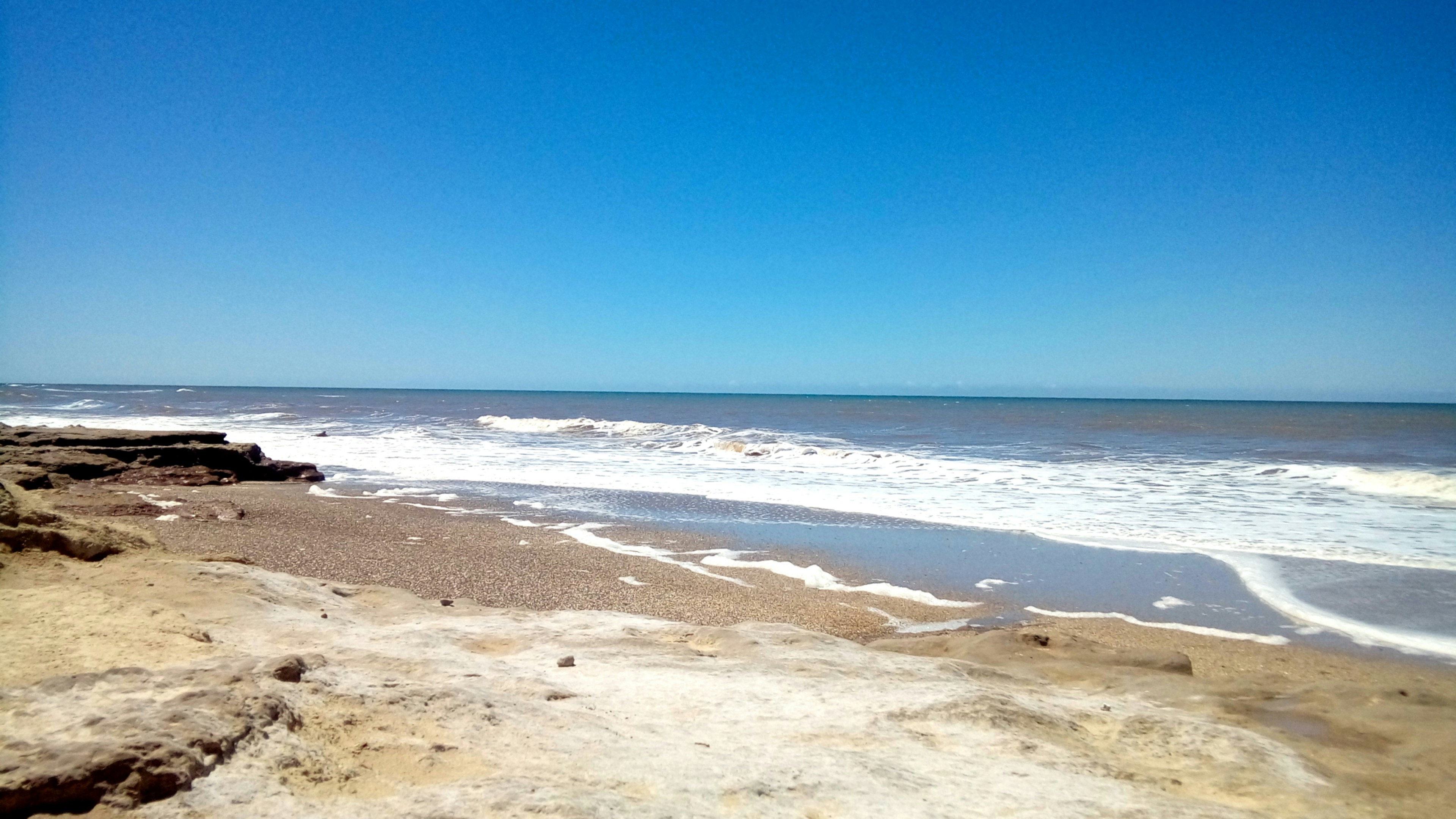 Waves crash on a rocky, sandy beach under clear sky.