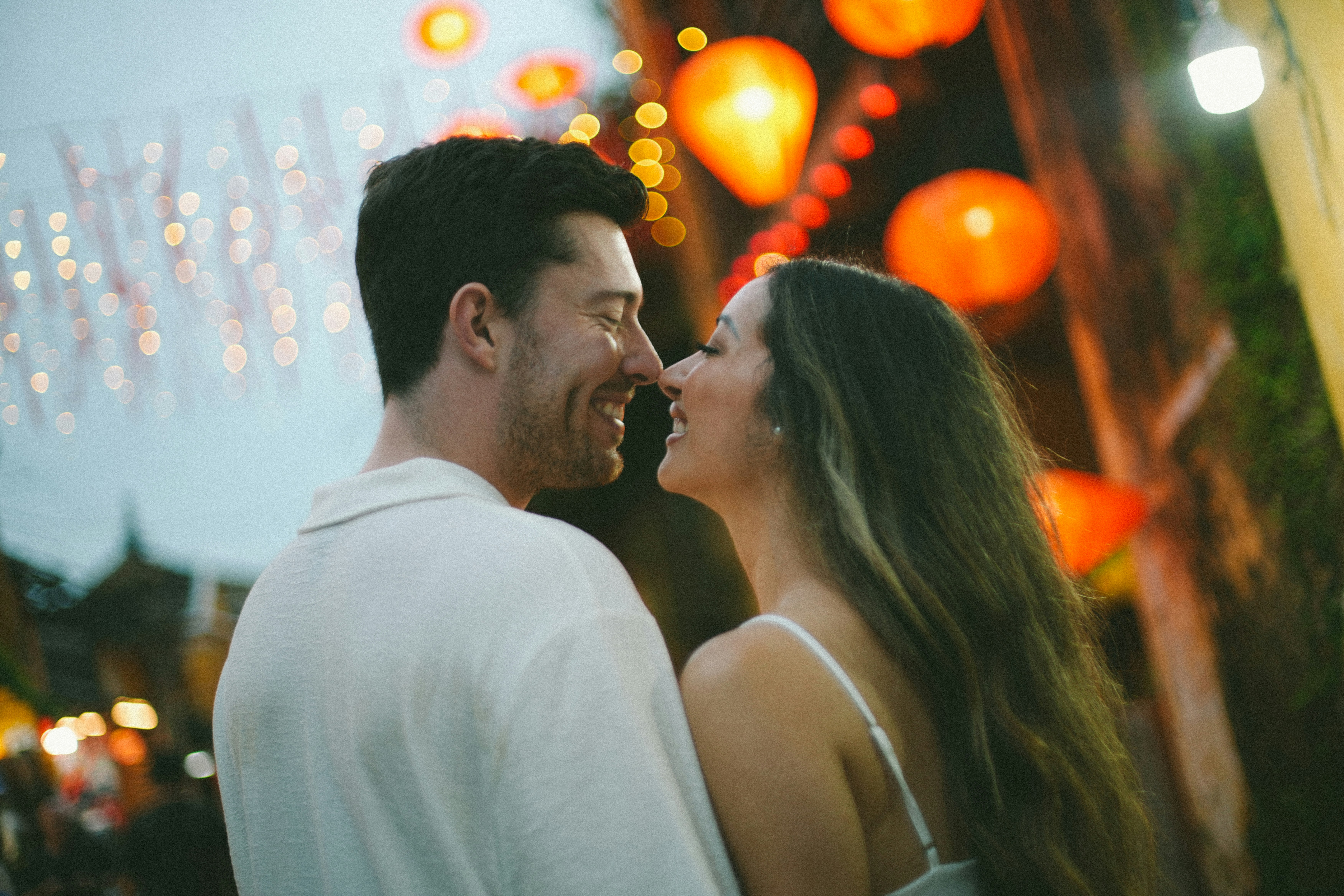 A couple smiles at each other with lanterns in background