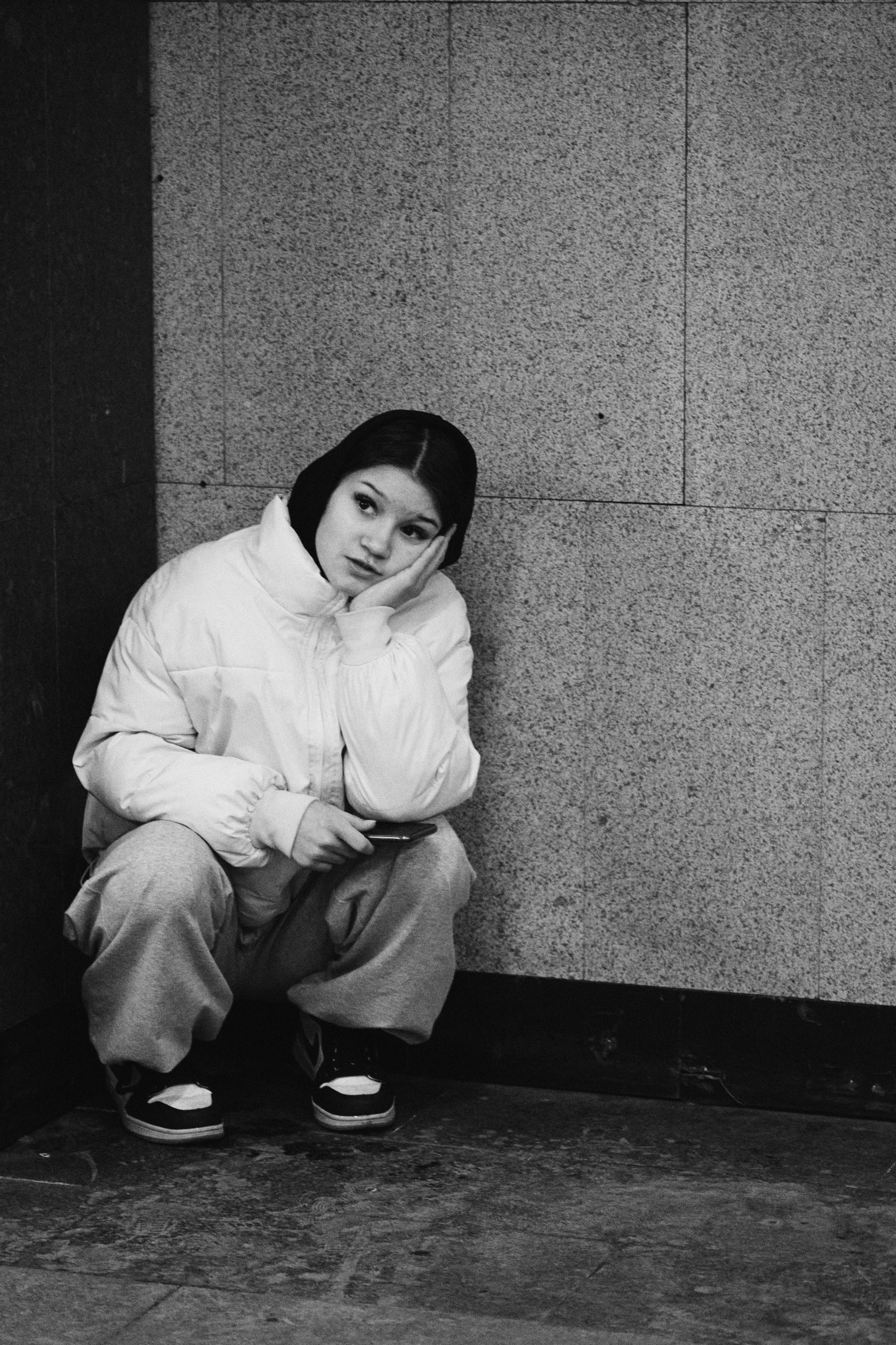 Young woman in winter jacket squats by wall.