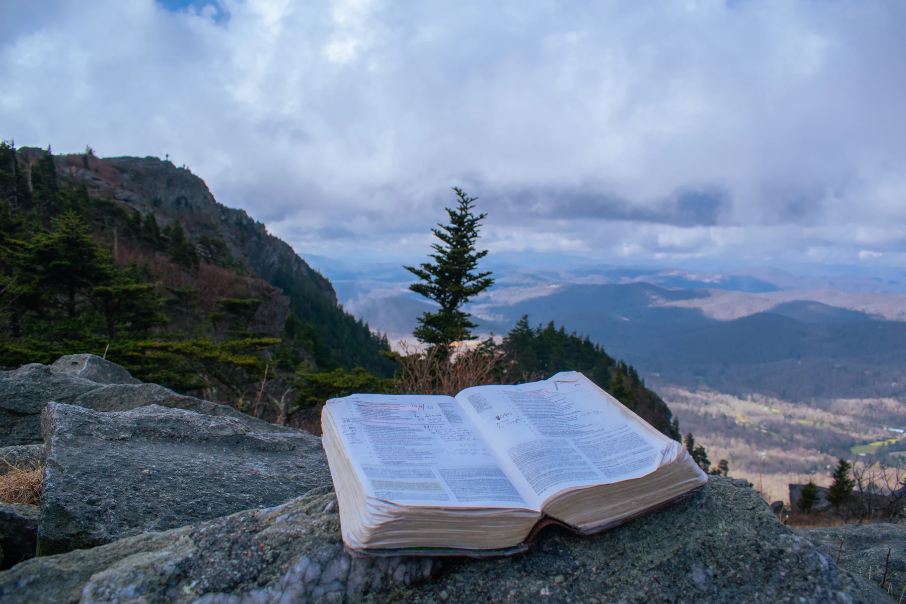 An open Bible resting on rocks with mountains behind
