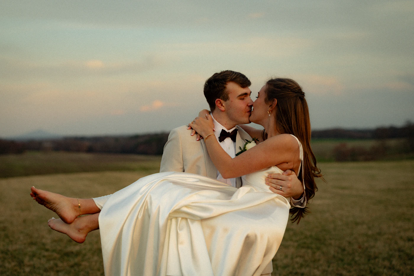 A groom carrying his bride in a romantic outdoor portrait at sunset with warm golden evening light illuminating the couple against an open landscape at their destination wedding