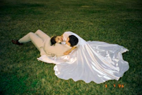 Newlyweds embrace while lying on grassy field.