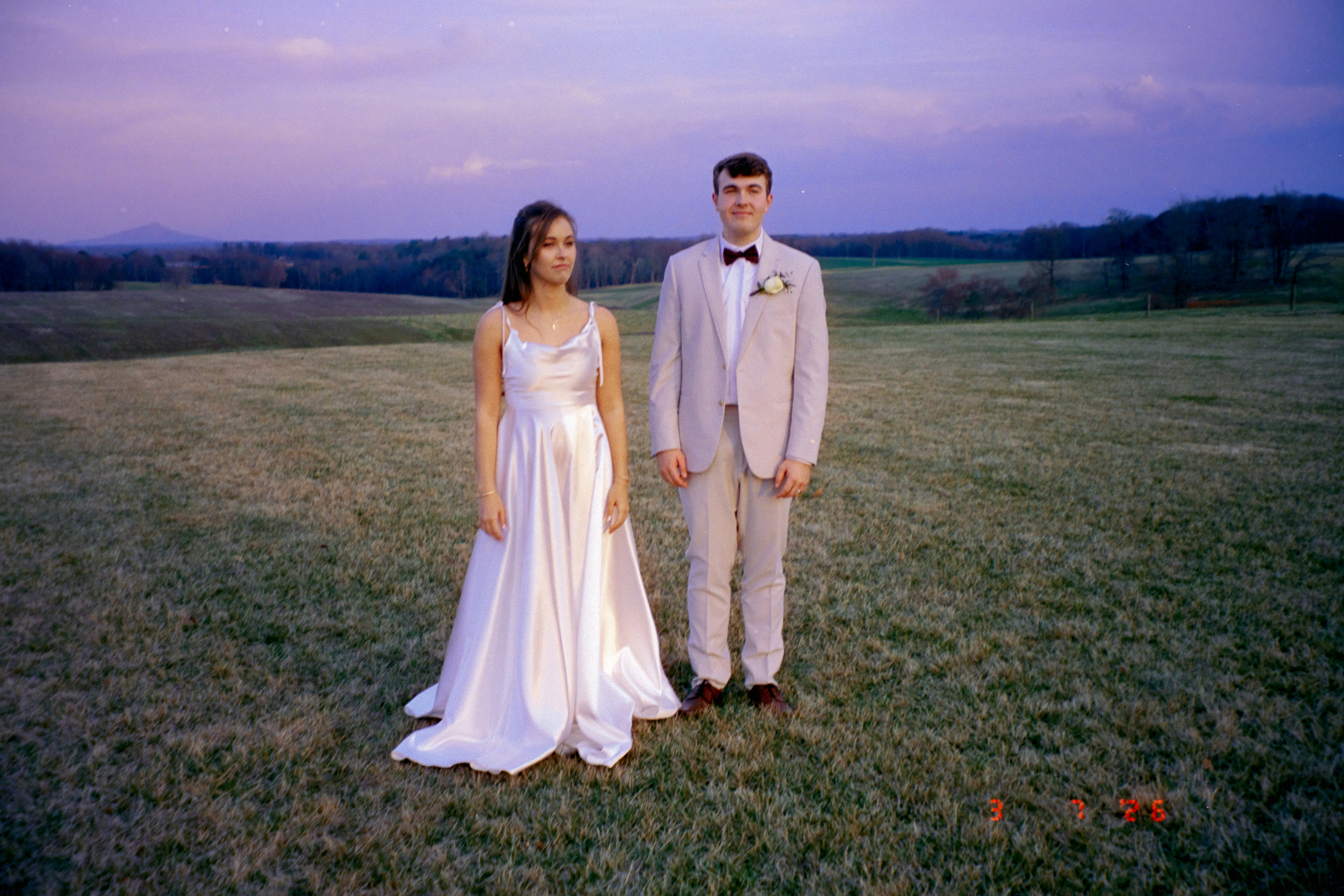 Jovem casal em traje formal parado em um campo gramado.