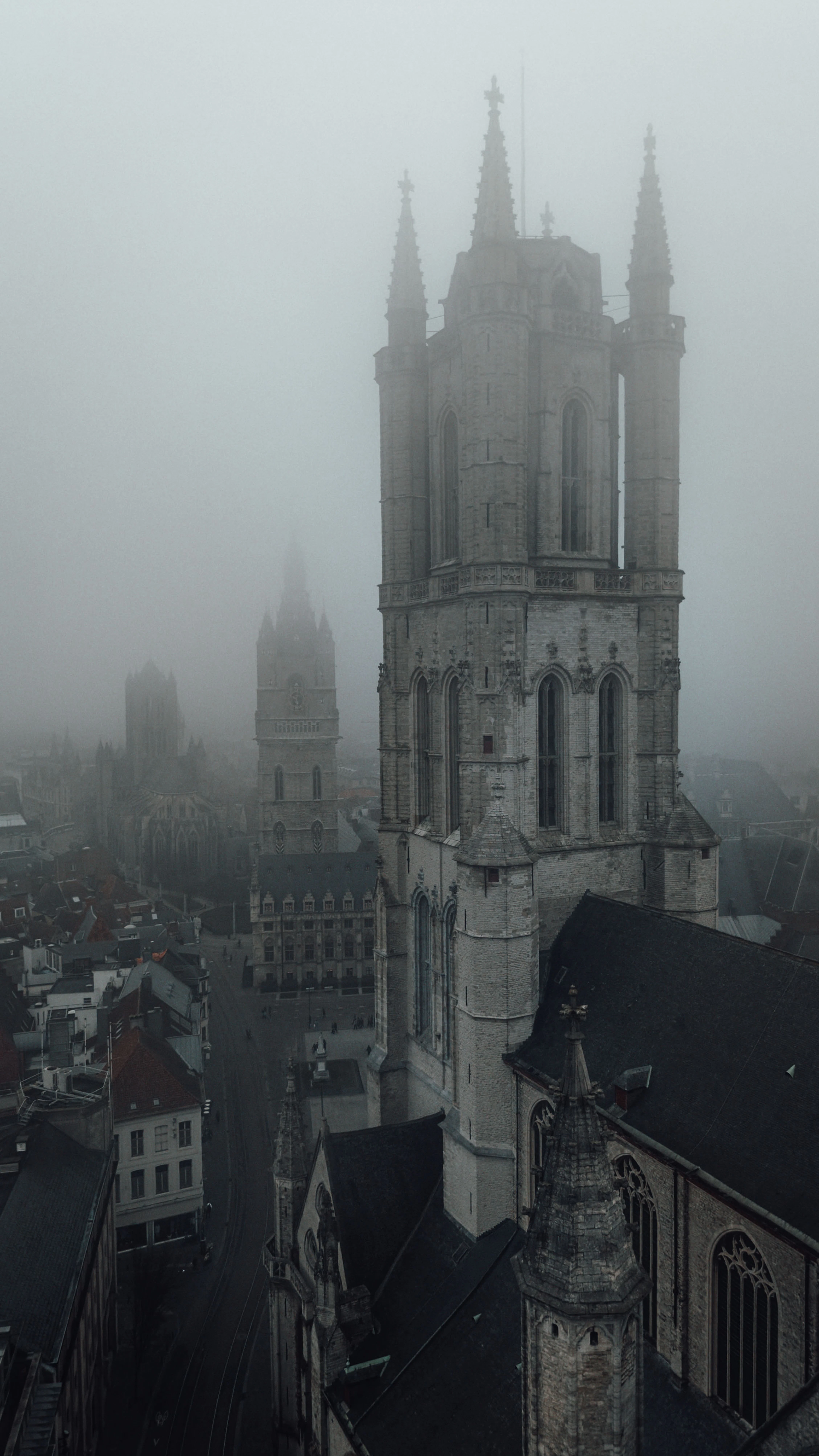 The Gothic tower of Saint Bavo's Cathedral in Ghent rising out of thick morning fog, with the Belfry a pale silhouette behind it and the empty square below.