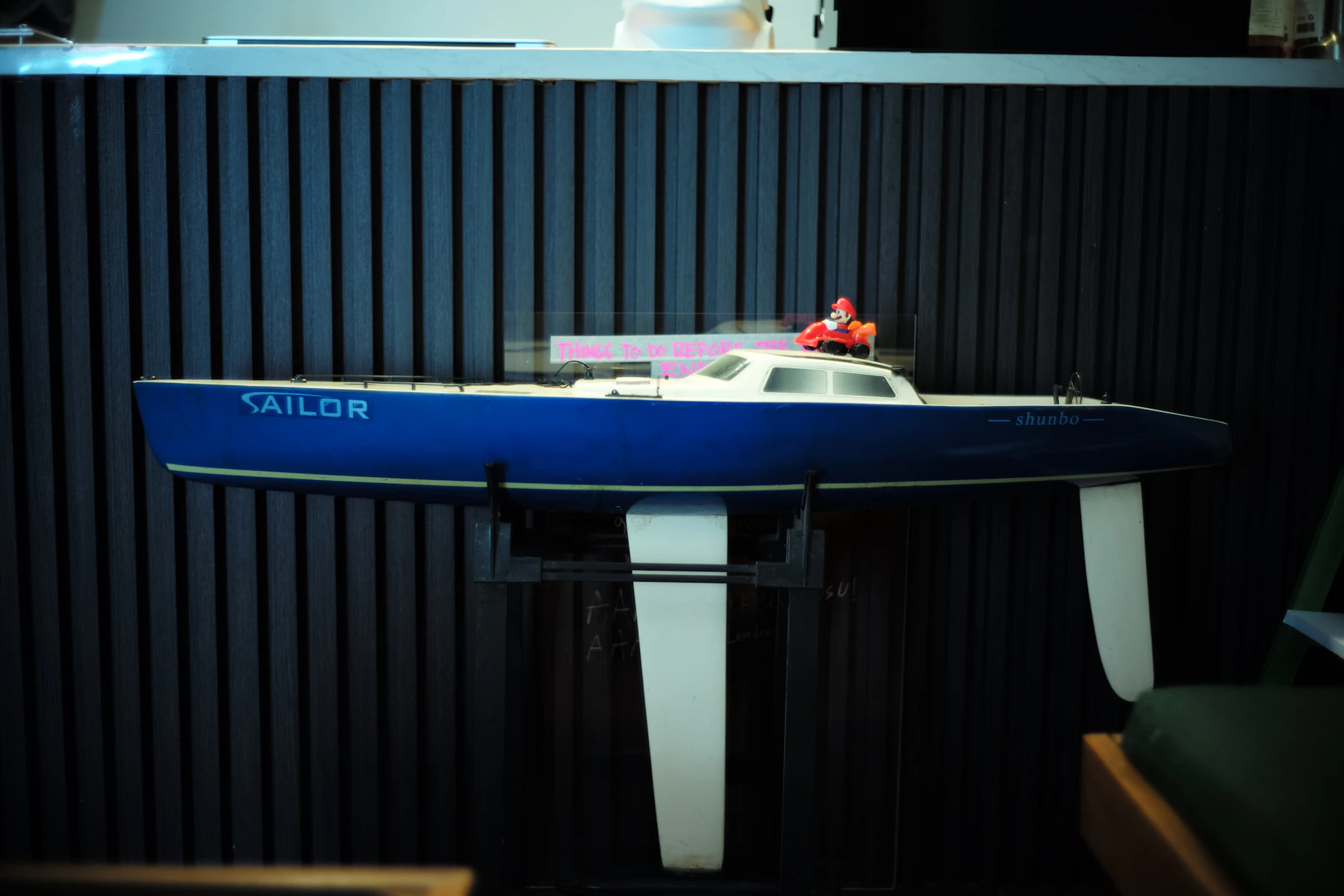 A blue model racing yacht on a display stand, its long white keel fin and rudder in view