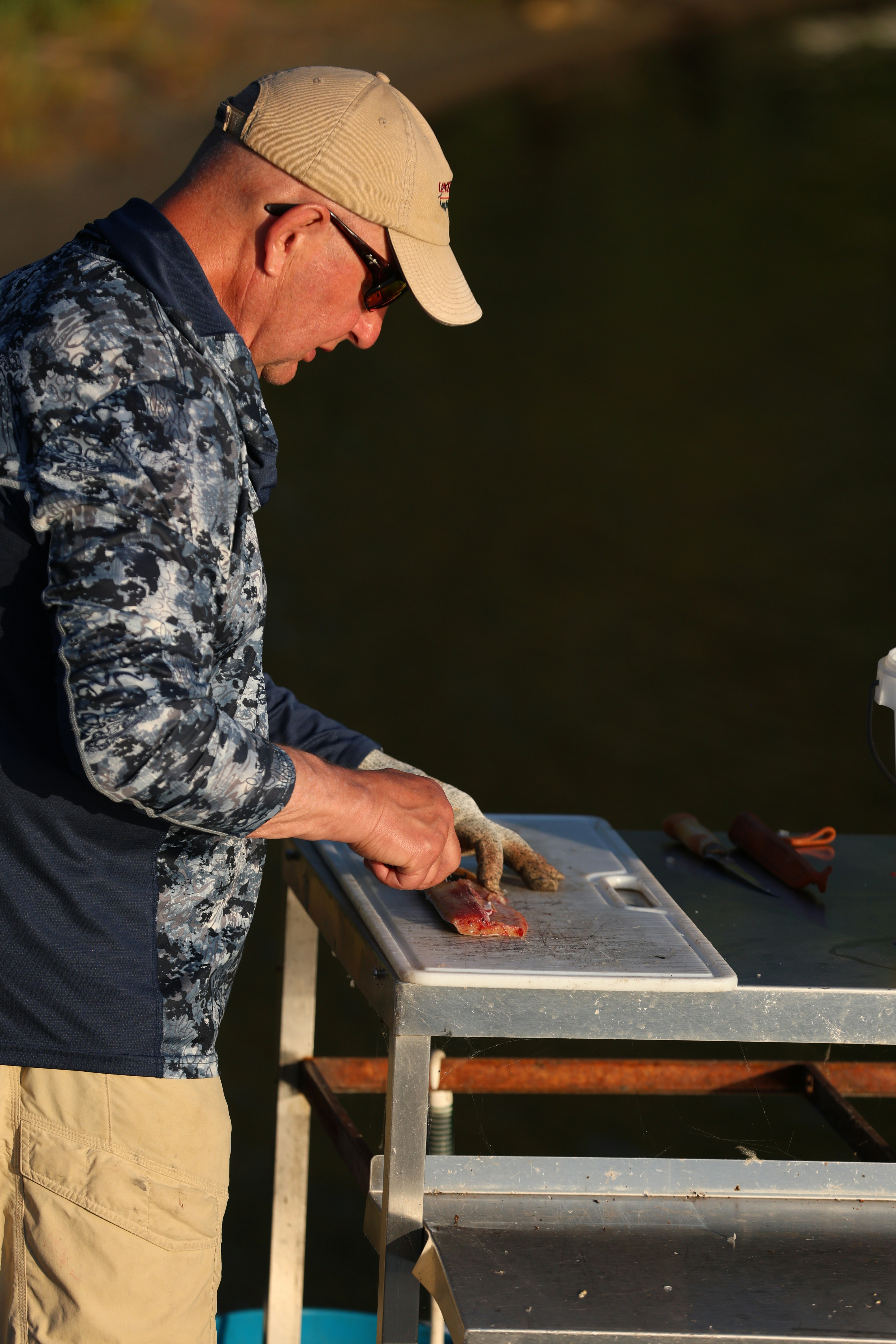 Man wearing a hat and sunglasses prepares fish outdoors.