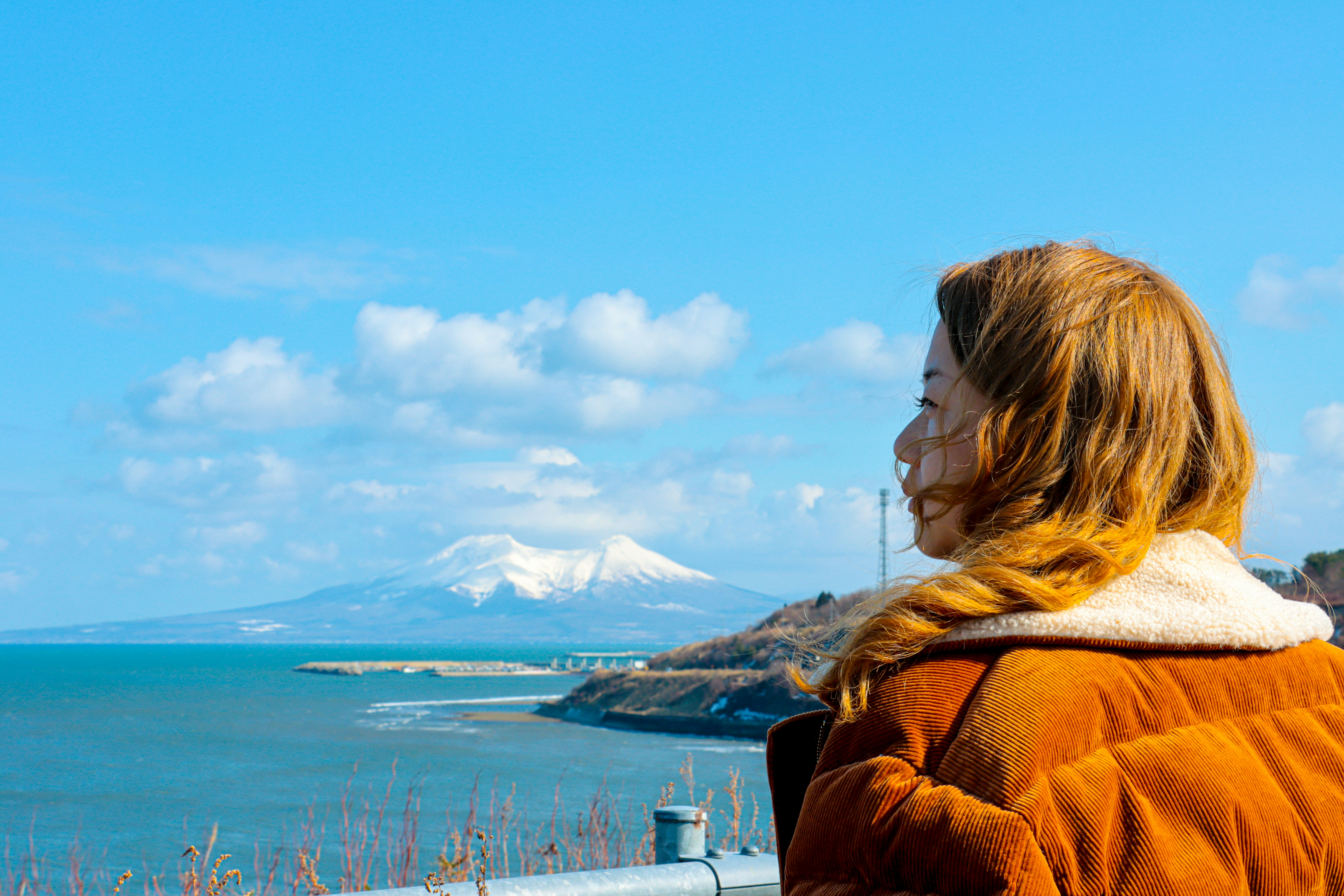 Woman in orange jacket looks at snow-capped mountain across ocean