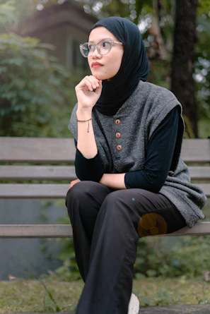 A woman wearing a hijab and glasses sits outdoors.