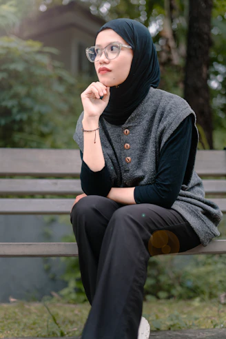 A woman wearing a hijab and glasses sits outdoors.