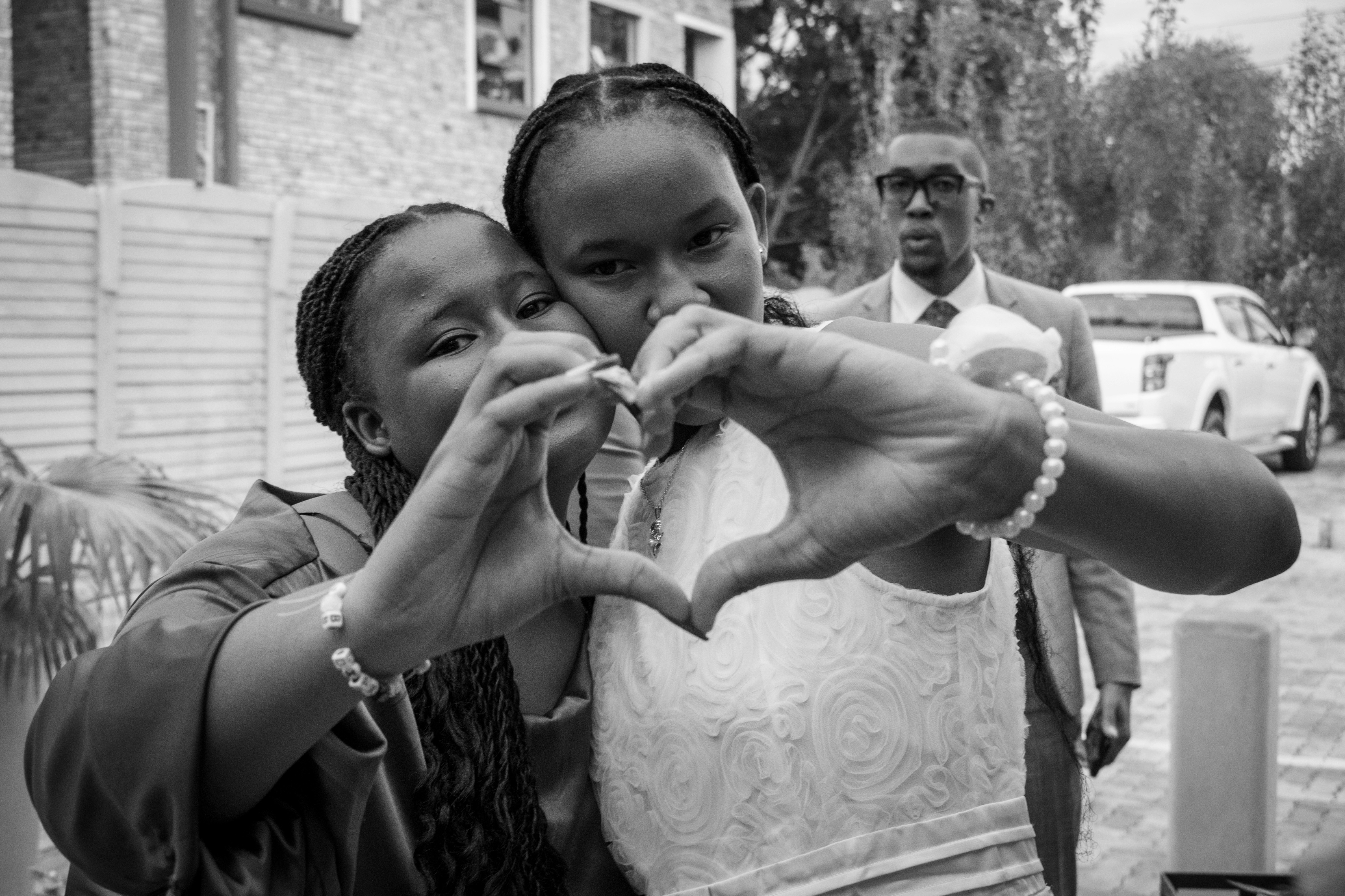 Two girls make heart shape with hands, man watches.
