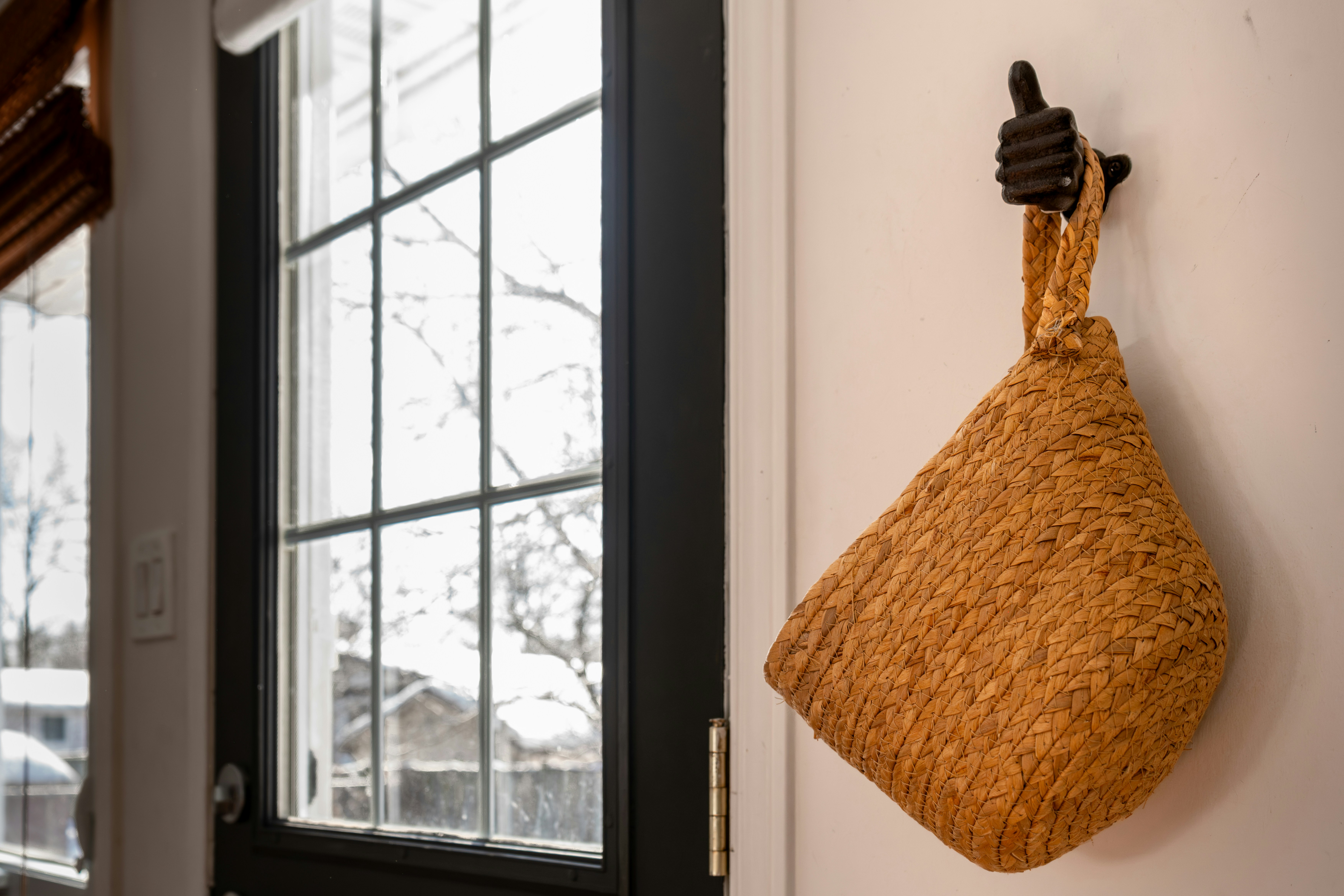 Woven straw bag hanging on a black cast iron hook beside a door with a window view, showing stylish door storage