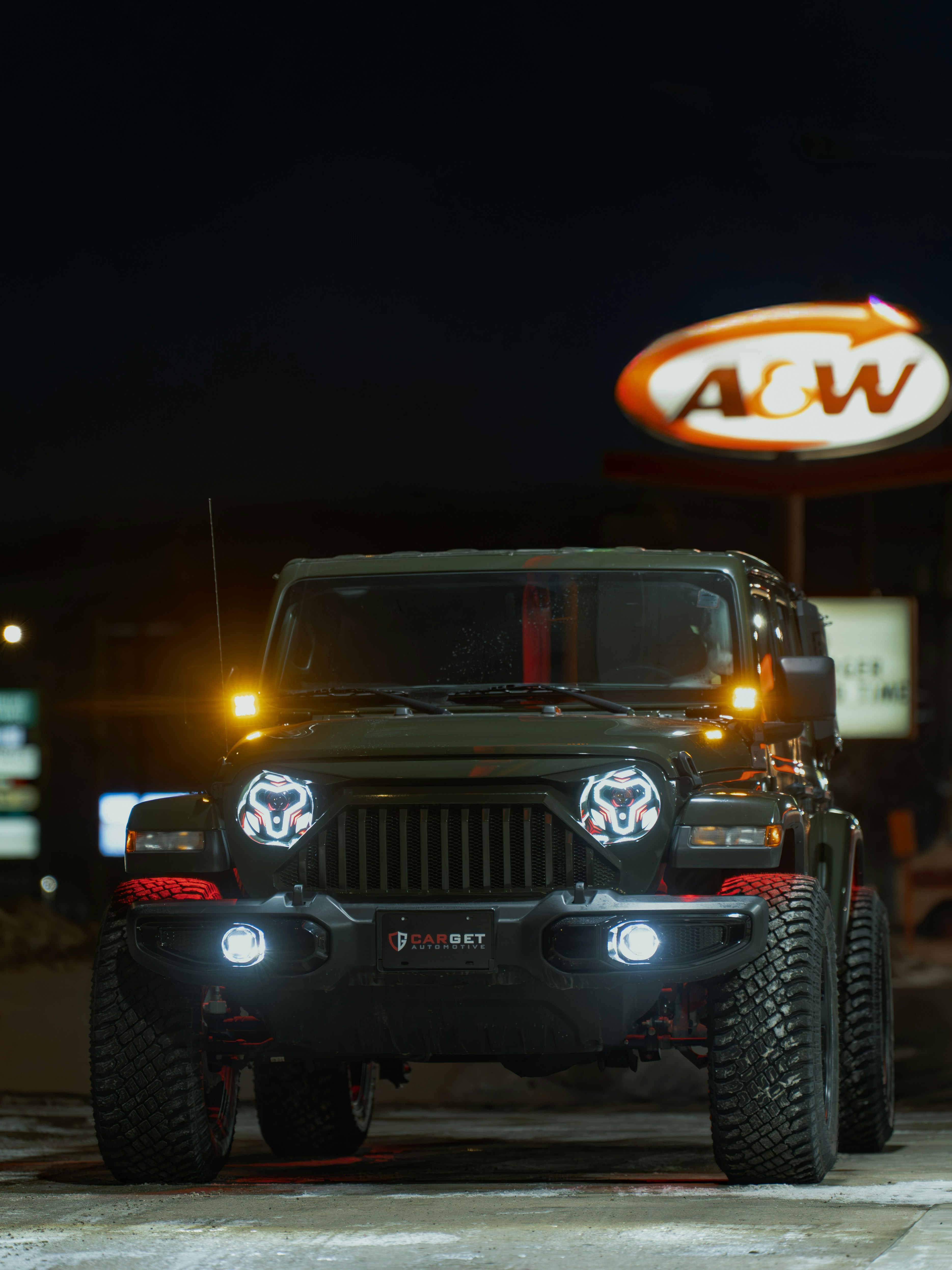 A dark green jeep wrangler parked at night near a&w.