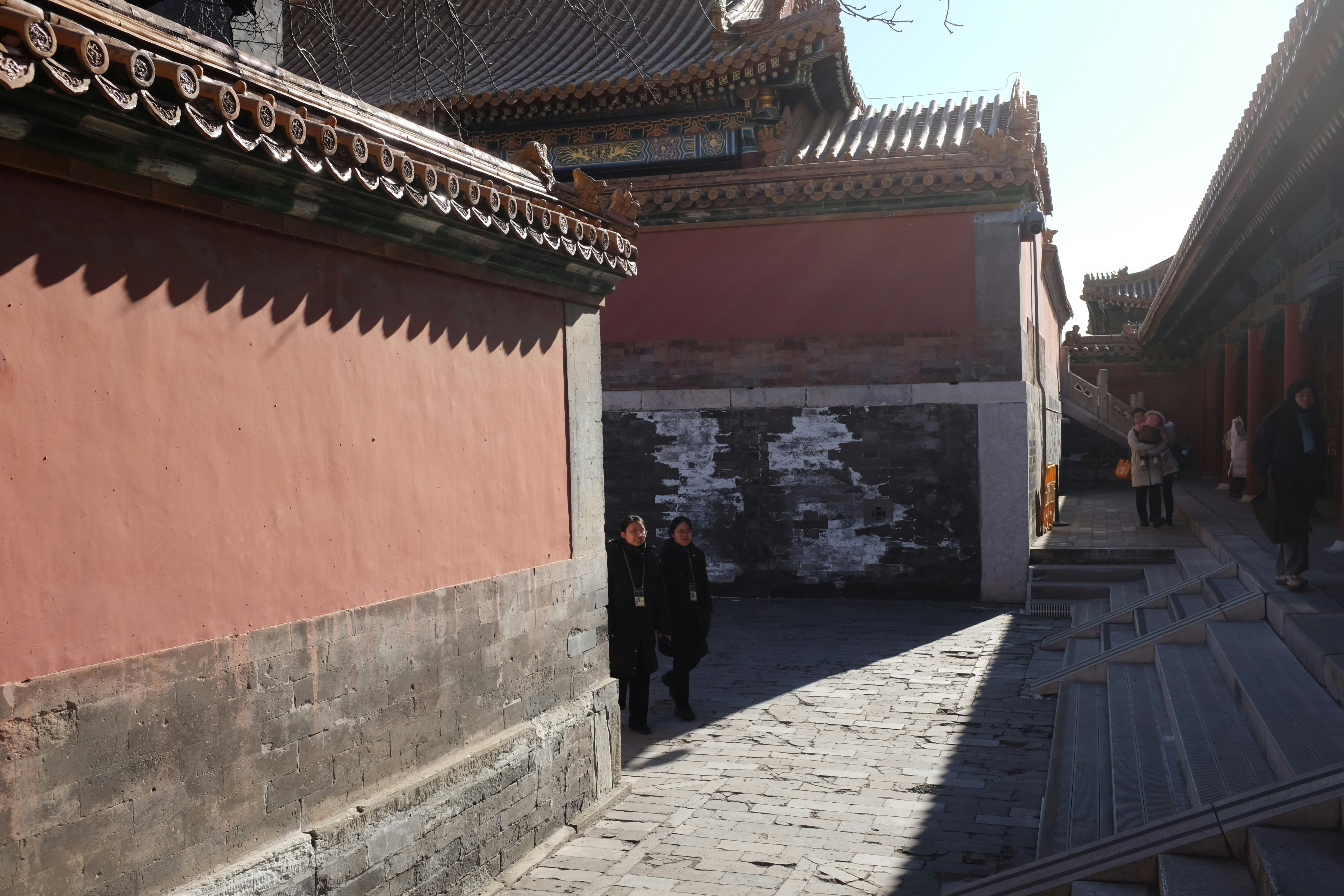 Two people walk down a sunlit alley between ancient buildings.