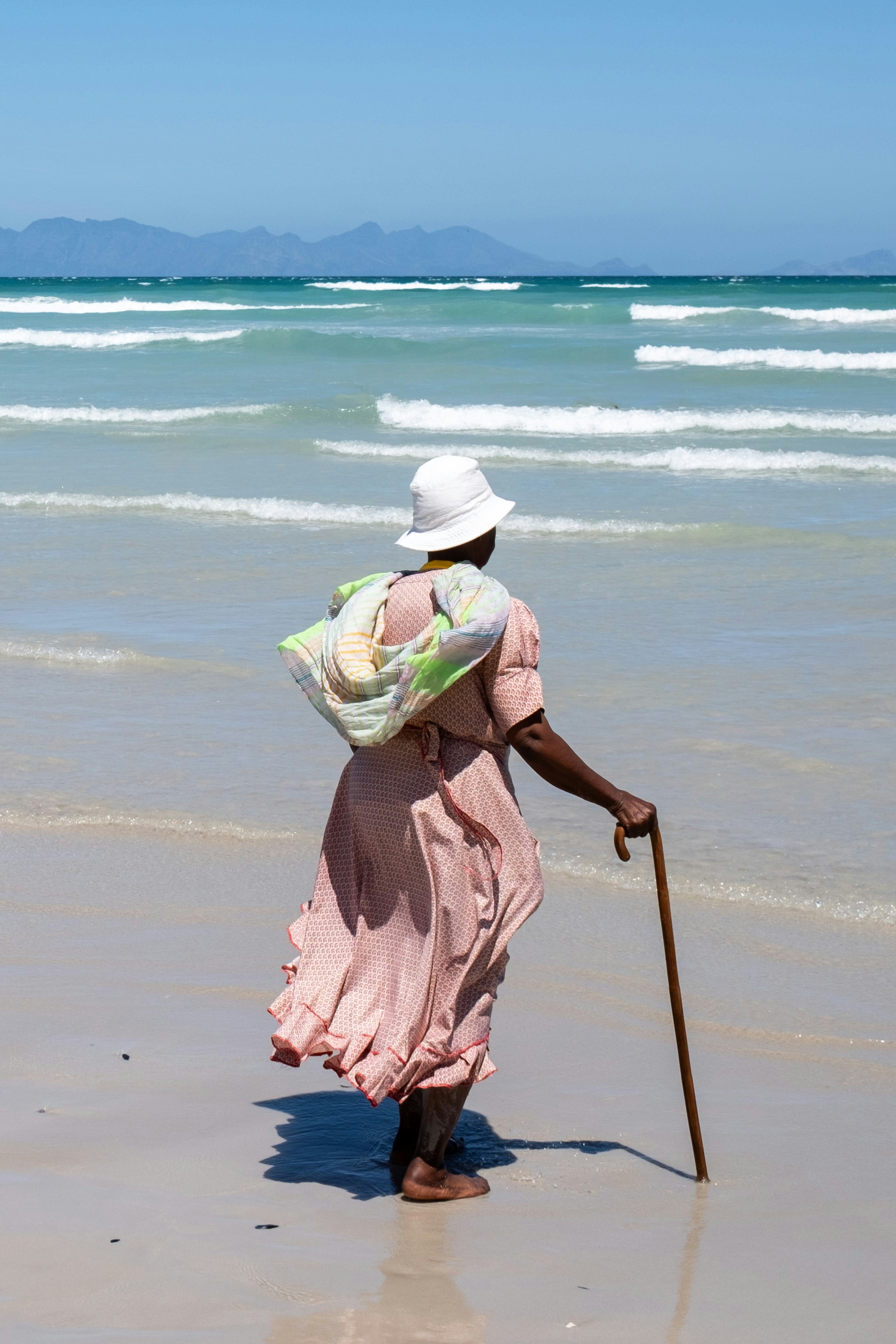 Elderly woman with a cane walks on a beach.