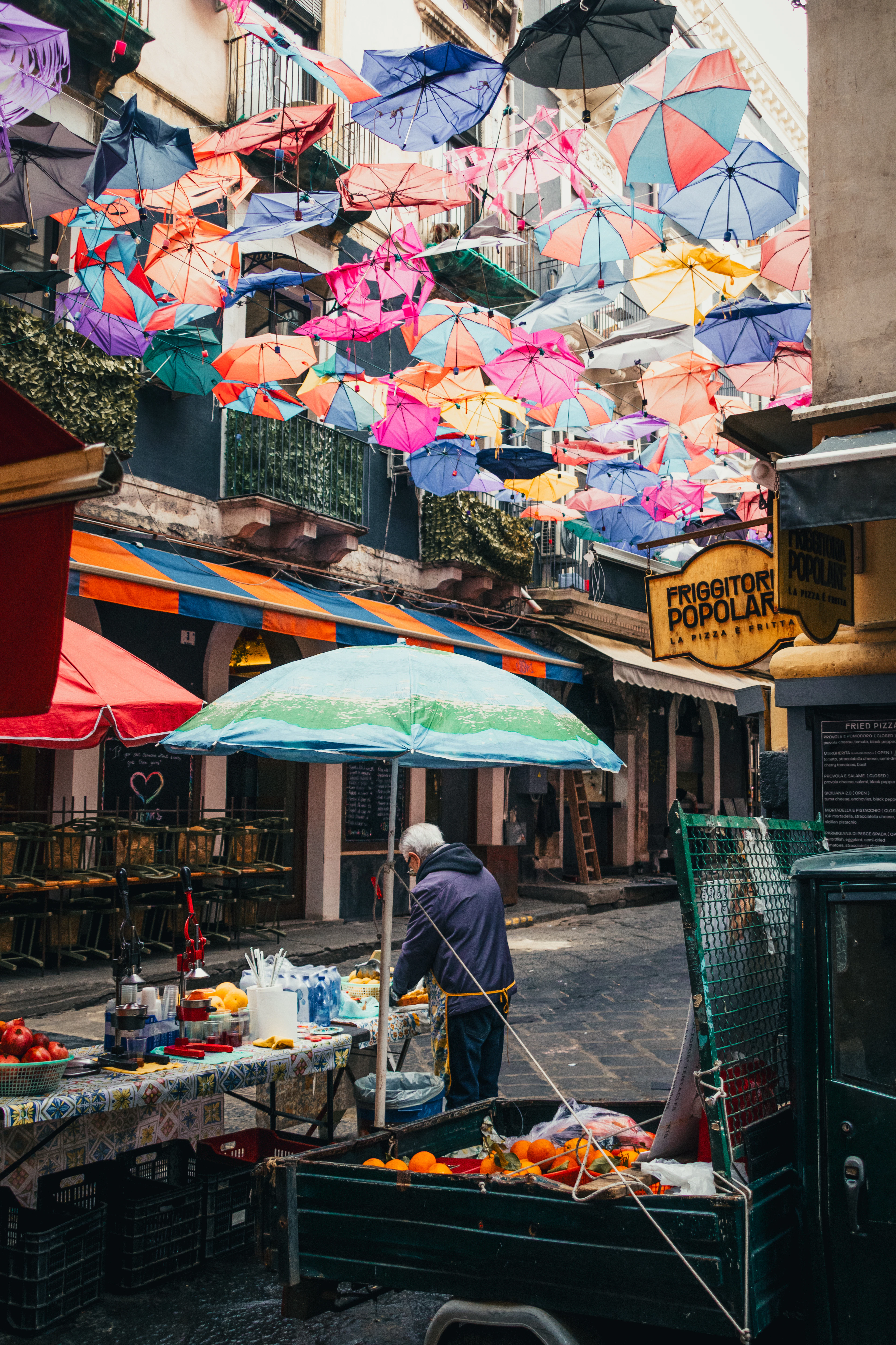 Colored umbrellas hang above a Catania alley where a vendor tends a citrus and fruit stall.