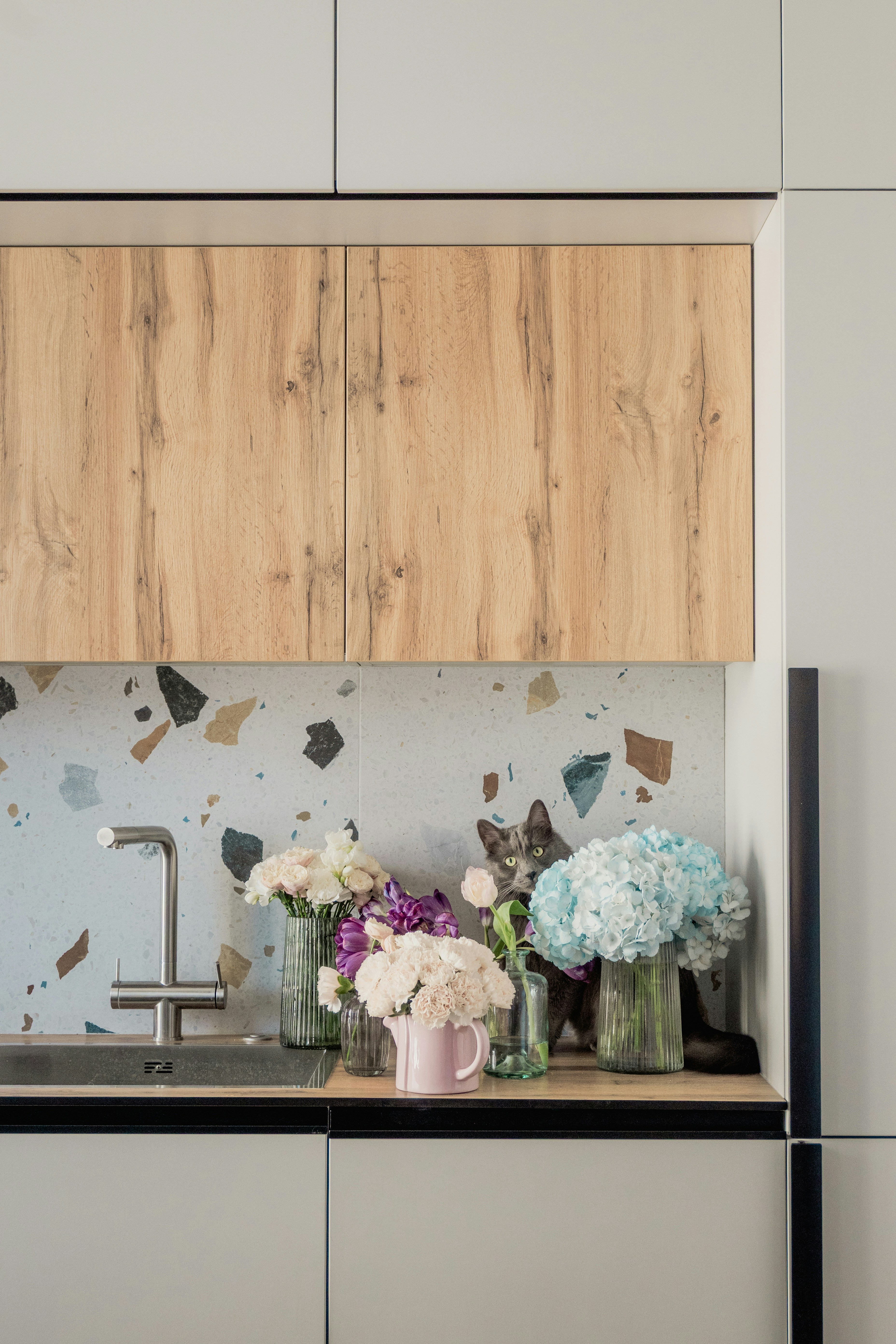 A cat peeks from behind flowers on a kitchen counter.
