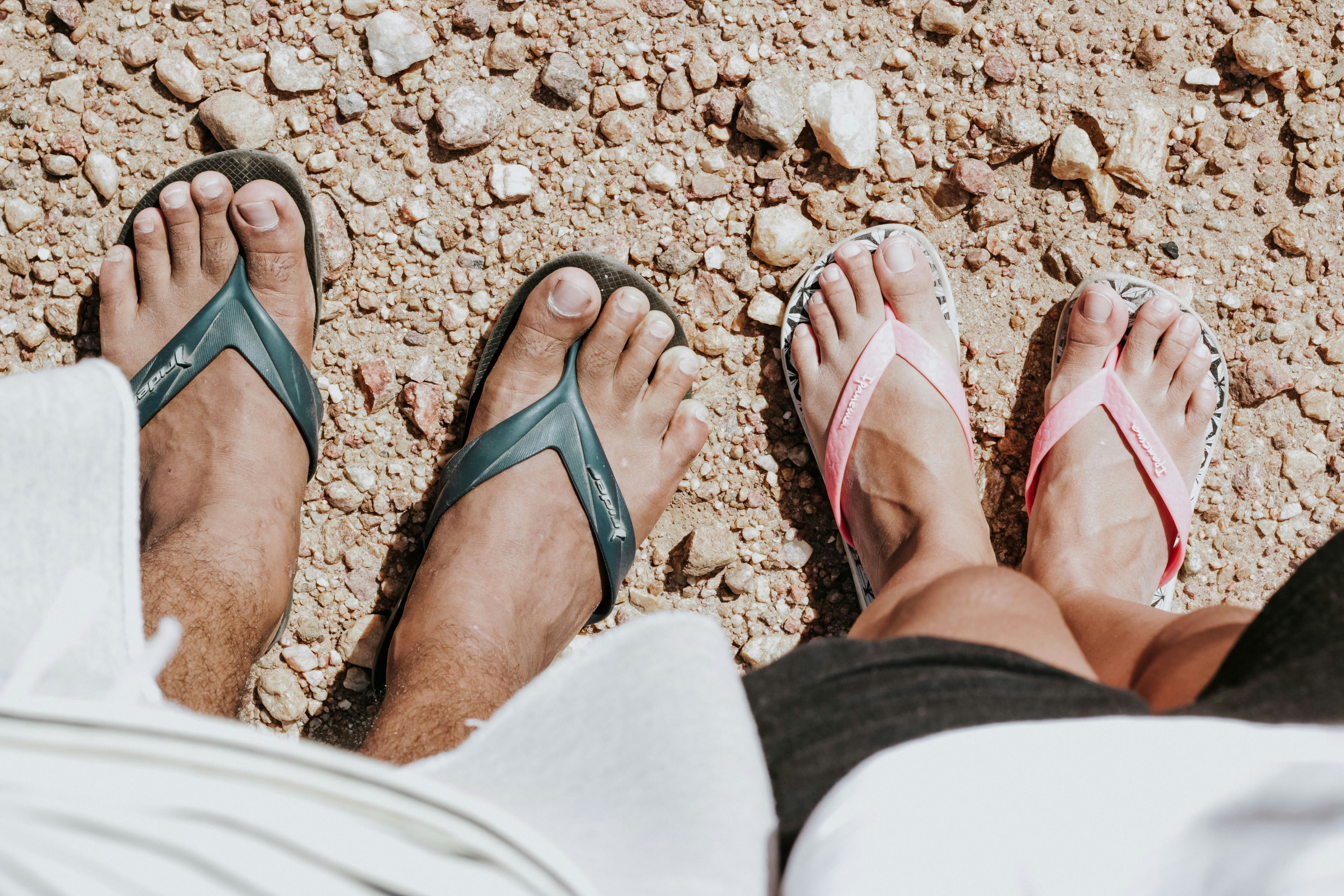Two pairs of feet wearing flip-flops on gravel.