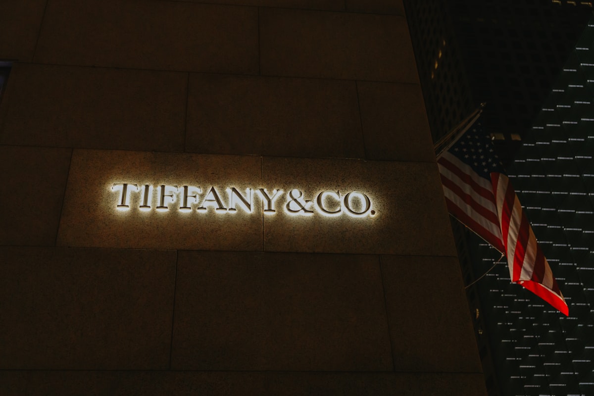 שלט TIFFANY & CO. מואר באותיות חרוטות על קיר אבן כהה בלילה, ודגל אמריקאי תלוי ממוט בפינת הבניין לצידו