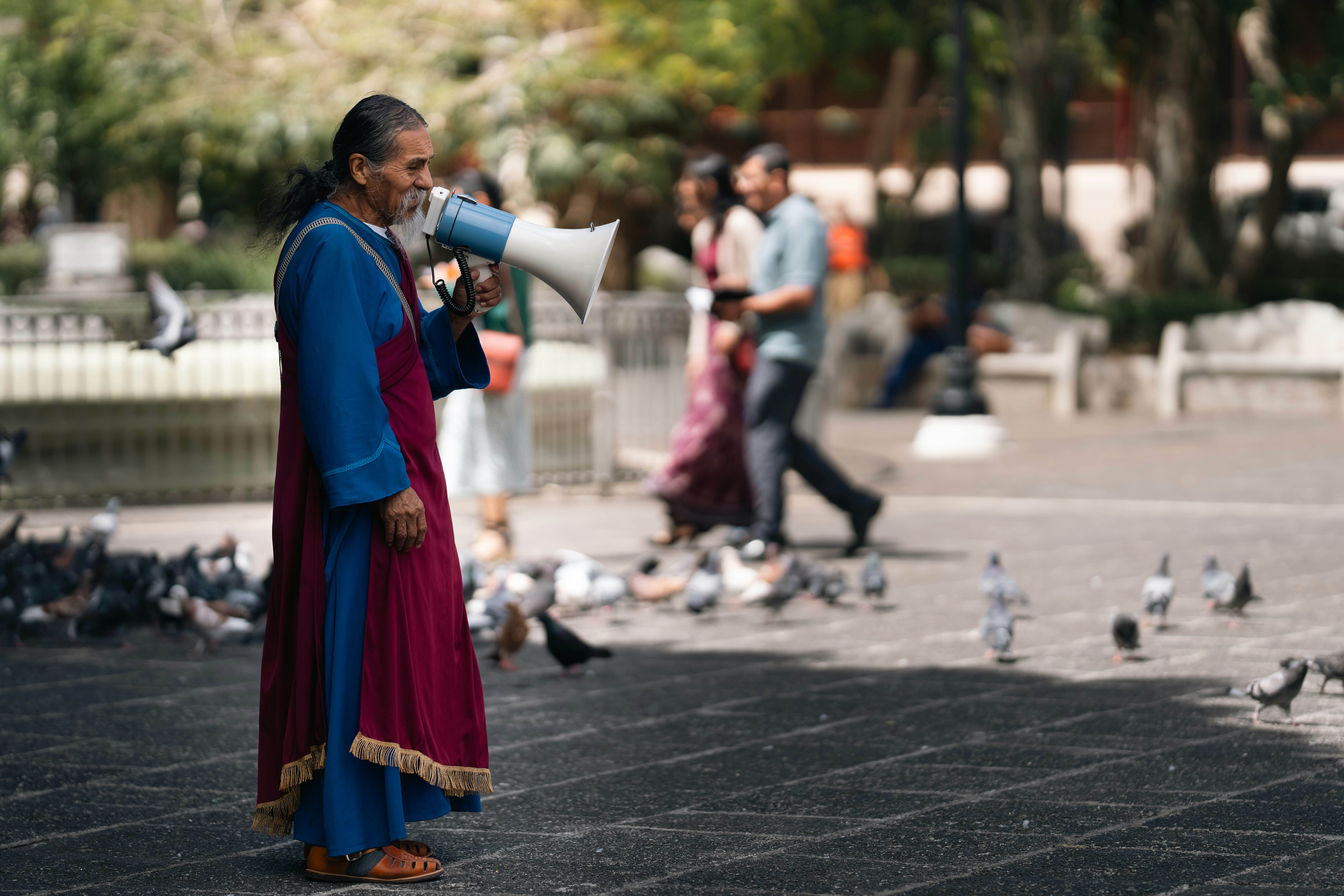 Elderly man with megaphone in park surrounded by pigeons.