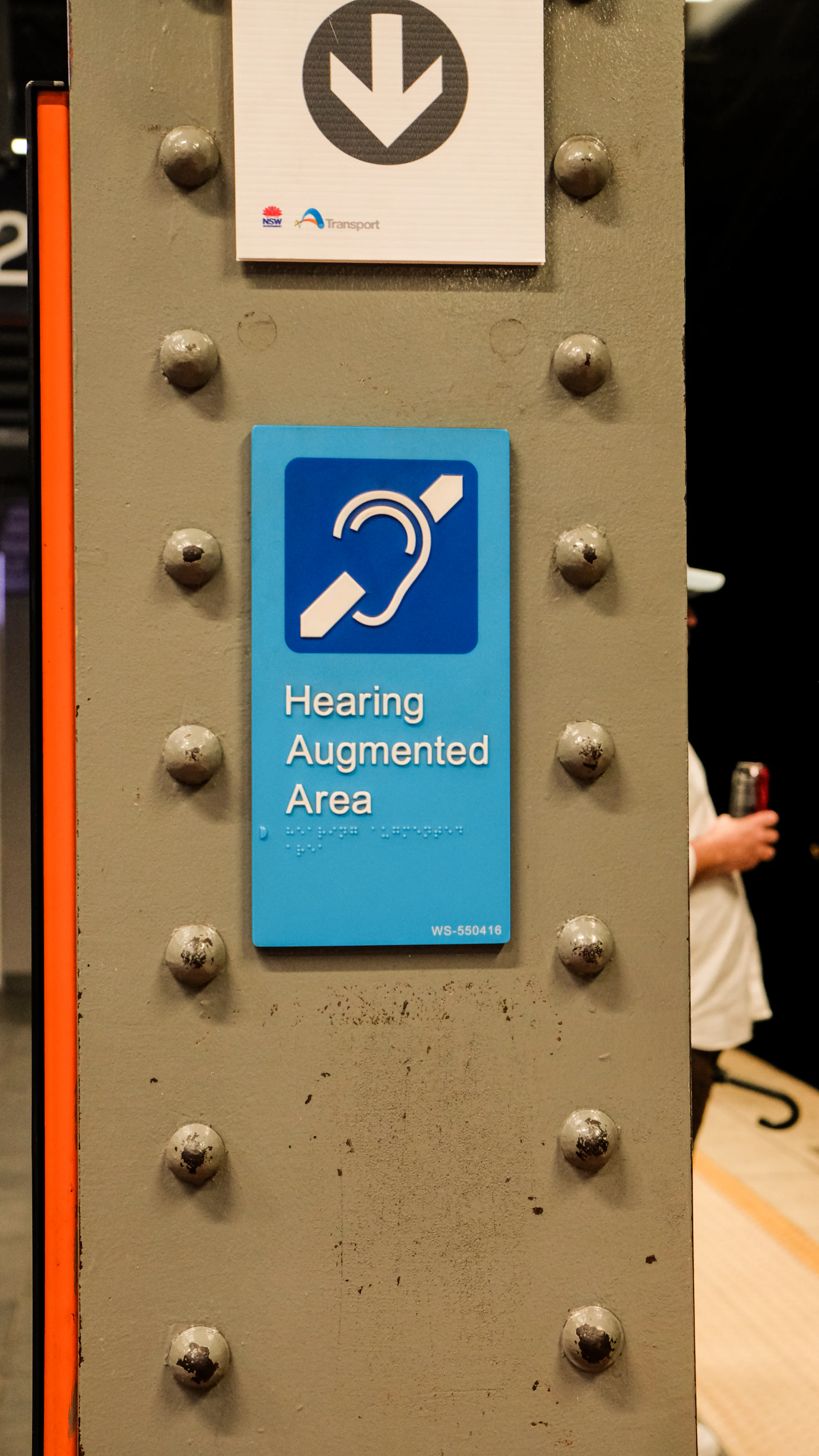 A blue sign on a station pillar reading Hearing Augmented Area, with a line of braille dots under the print