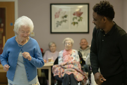 Elderly woman dances with caregiver in a community room.