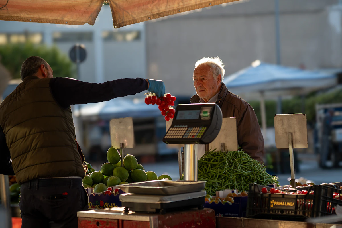 A market trader holding up a bunch of tomatoes over a set of scales while an older customer weighs up the offer