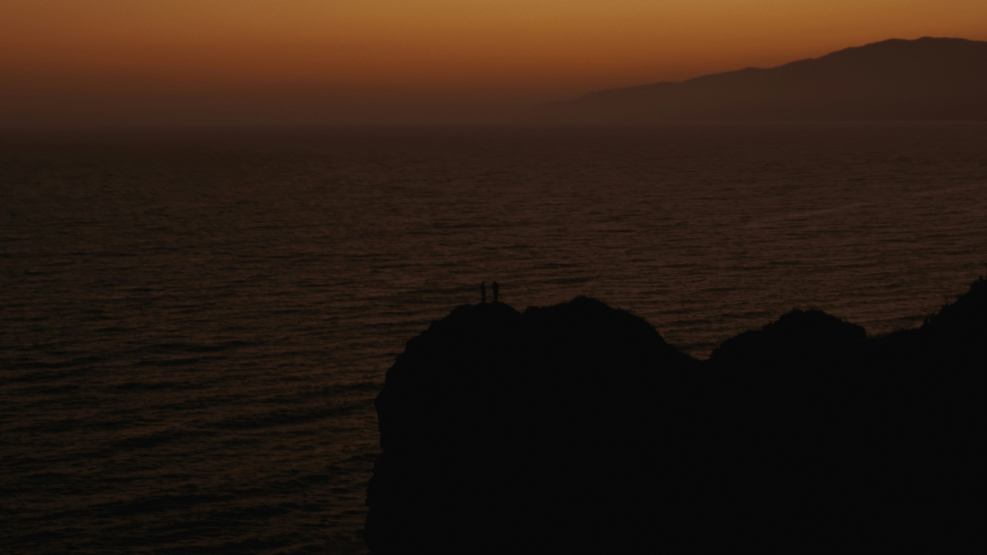シルエットの人物が夕暮れの海を見下ろす崖に立っている